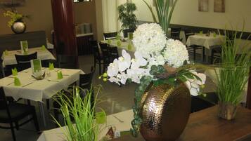 A restaurant interior with tables set for dining, featuring decorative flower arrangements and green plants.