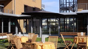 An outdoor seating area with wooden chairs and tables, a covered patio, and a building with multiple floors and balconies. - VCH Hotel Greifswald (Greifswald)