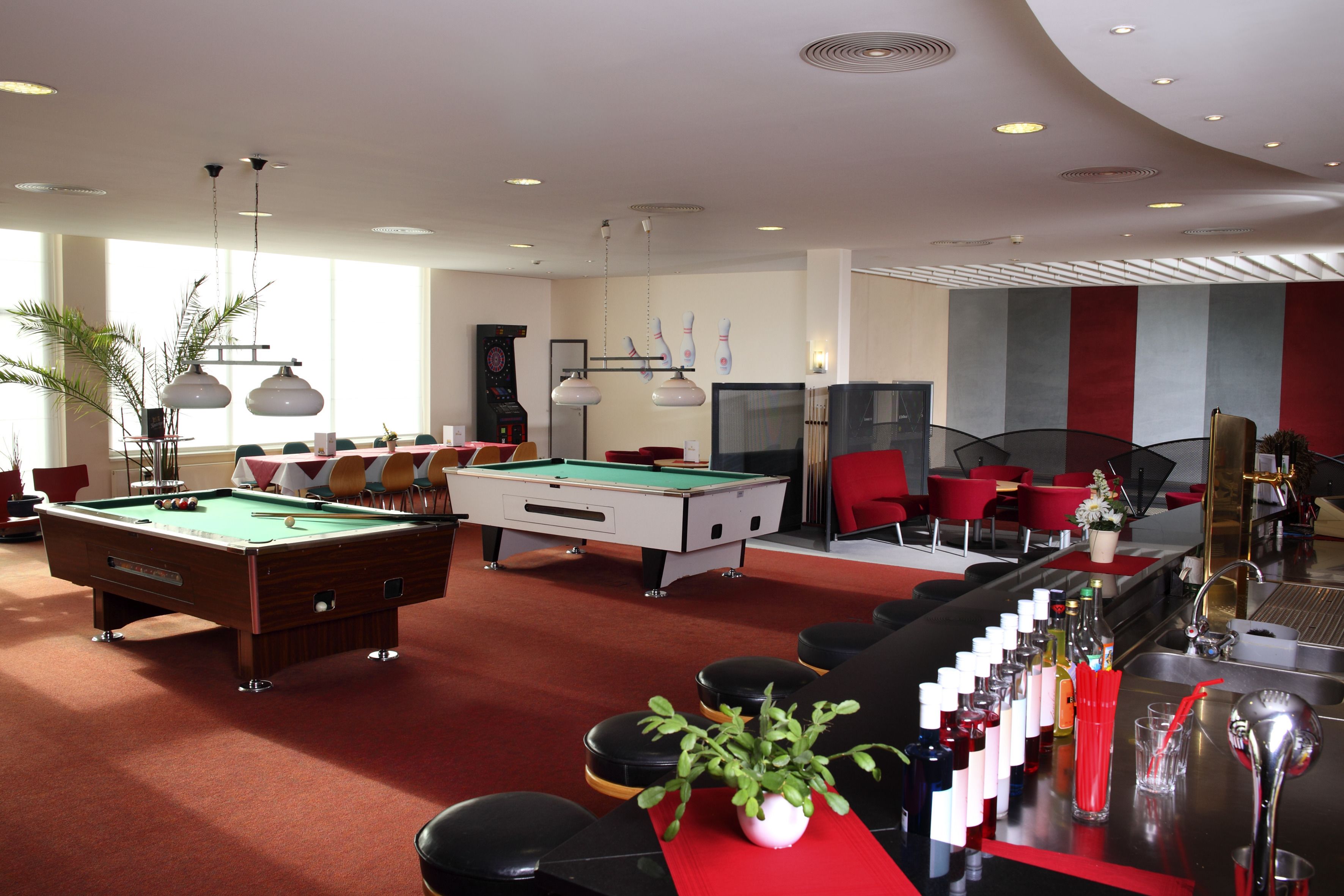 A pool table, bar area with stools, and red chairs in a well-lit room with large windows.