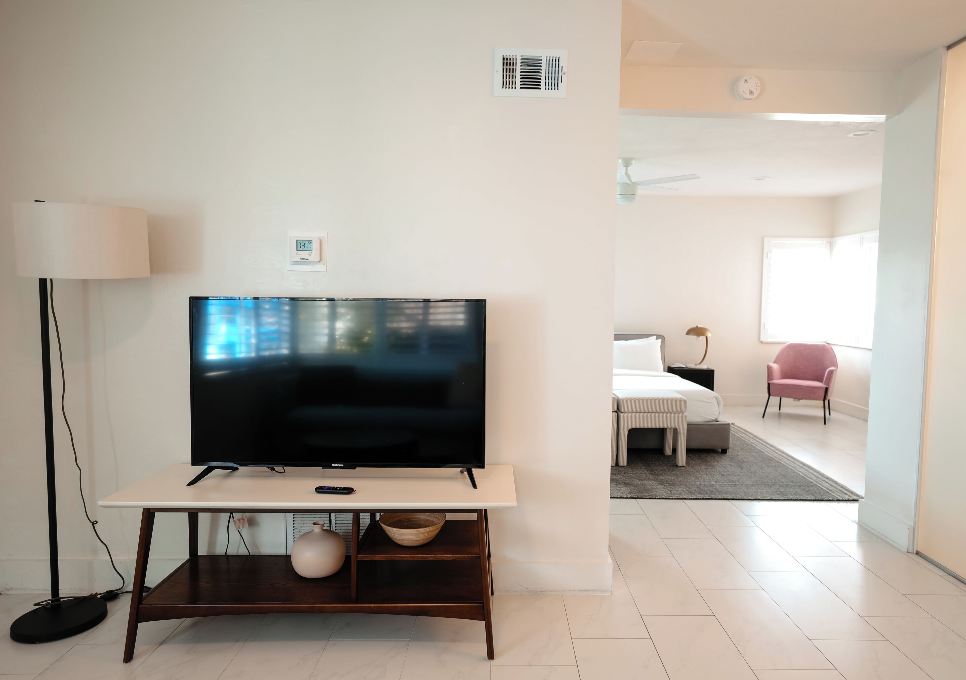 A modern living room with a flat-screen TV on a wooden stand, a lamp, and a thermostat on the wall.