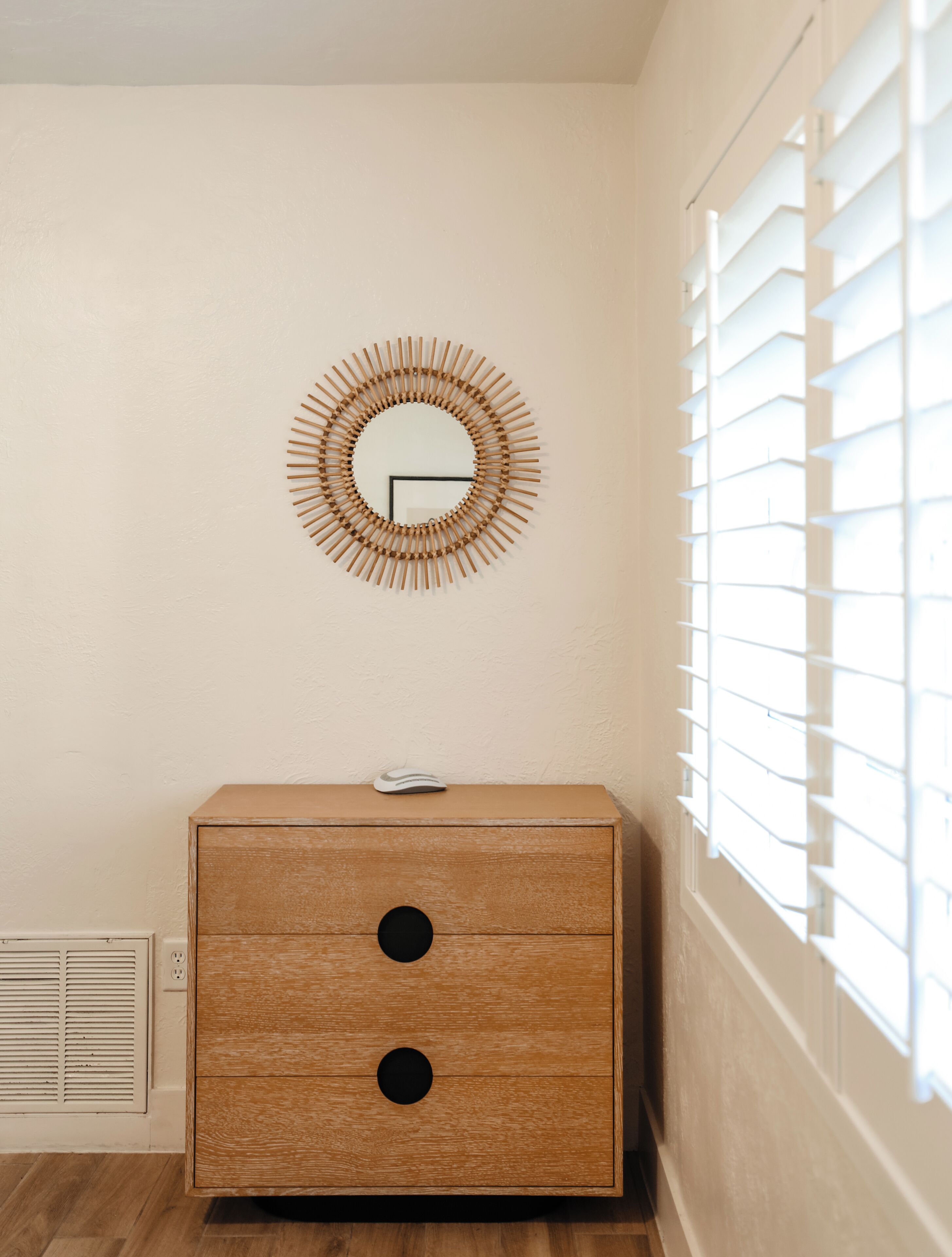 A room with a wooden dresser, a wall-mounted mirror, and a window with blinds.