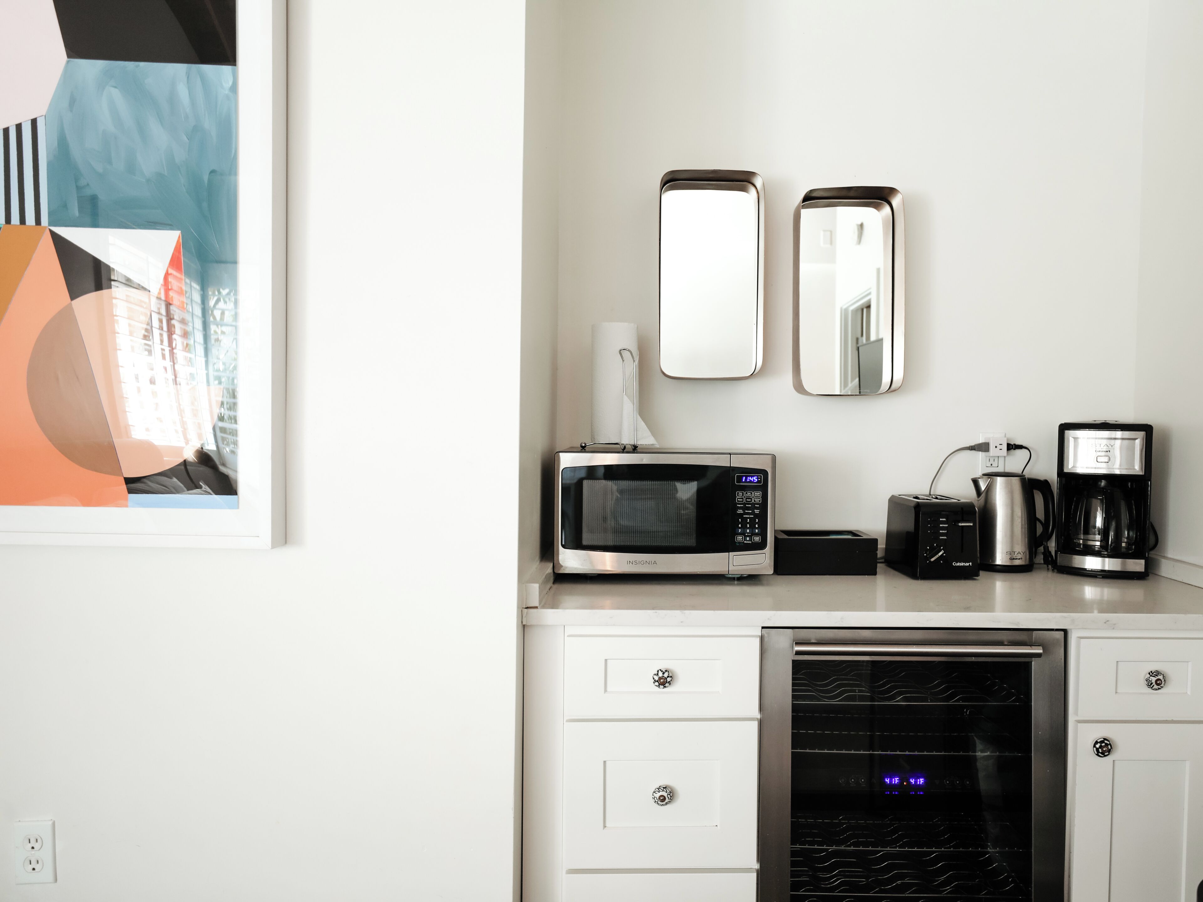 A modern kitchen with a microwave, coffee maker, and oven. There are two wall mirrors and a framed artwork above the counter.