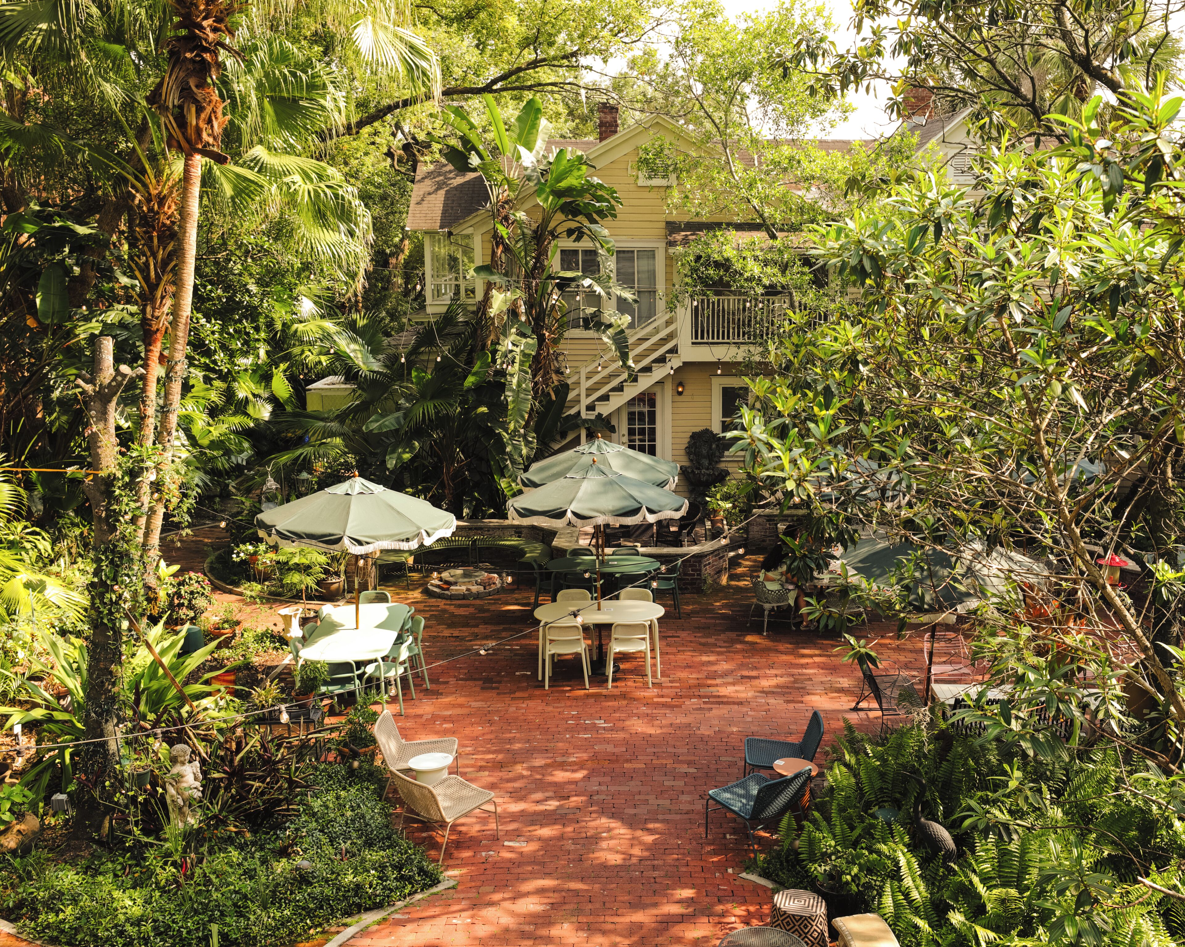 A patio with tables and chairs, surrounded by lush greenery and a house in the background.