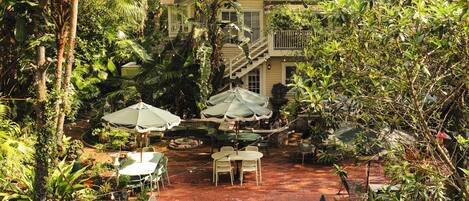 A patio with tables and chairs, surrounded by lush greenery and a house in the background.