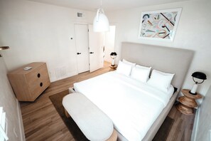 A modern bedroom with a large bed, a bedside table, a wooden dresser, and a hanging light fixture.