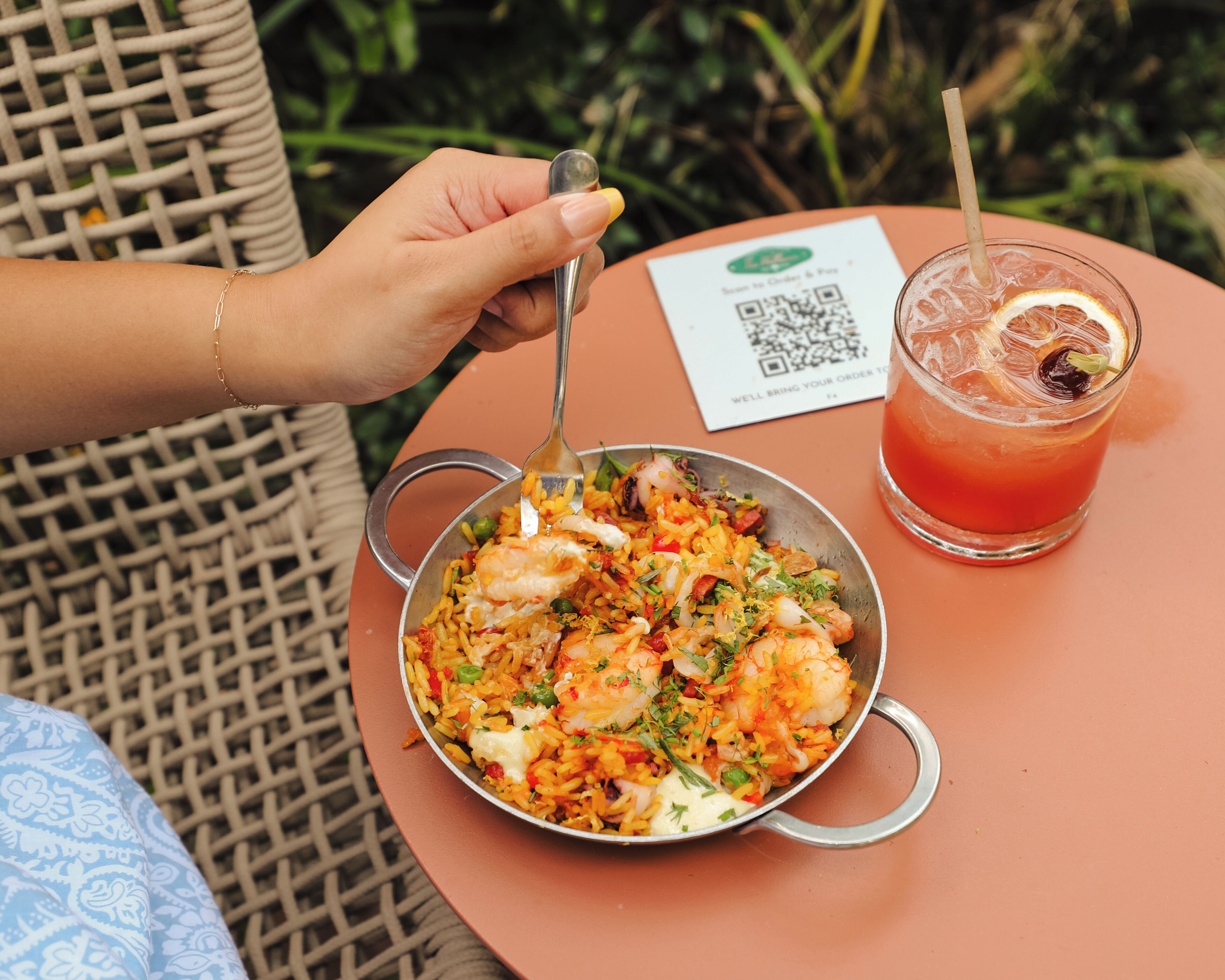 A person dining outdoors with a bowl of shrimp and rice, a glass of red drink, and a QR code on a table.