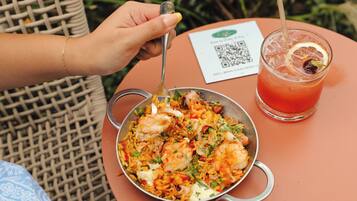 A person dining outdoors with a bowl of shrimp and rice, a glass of red drink, and a QR code on a table.