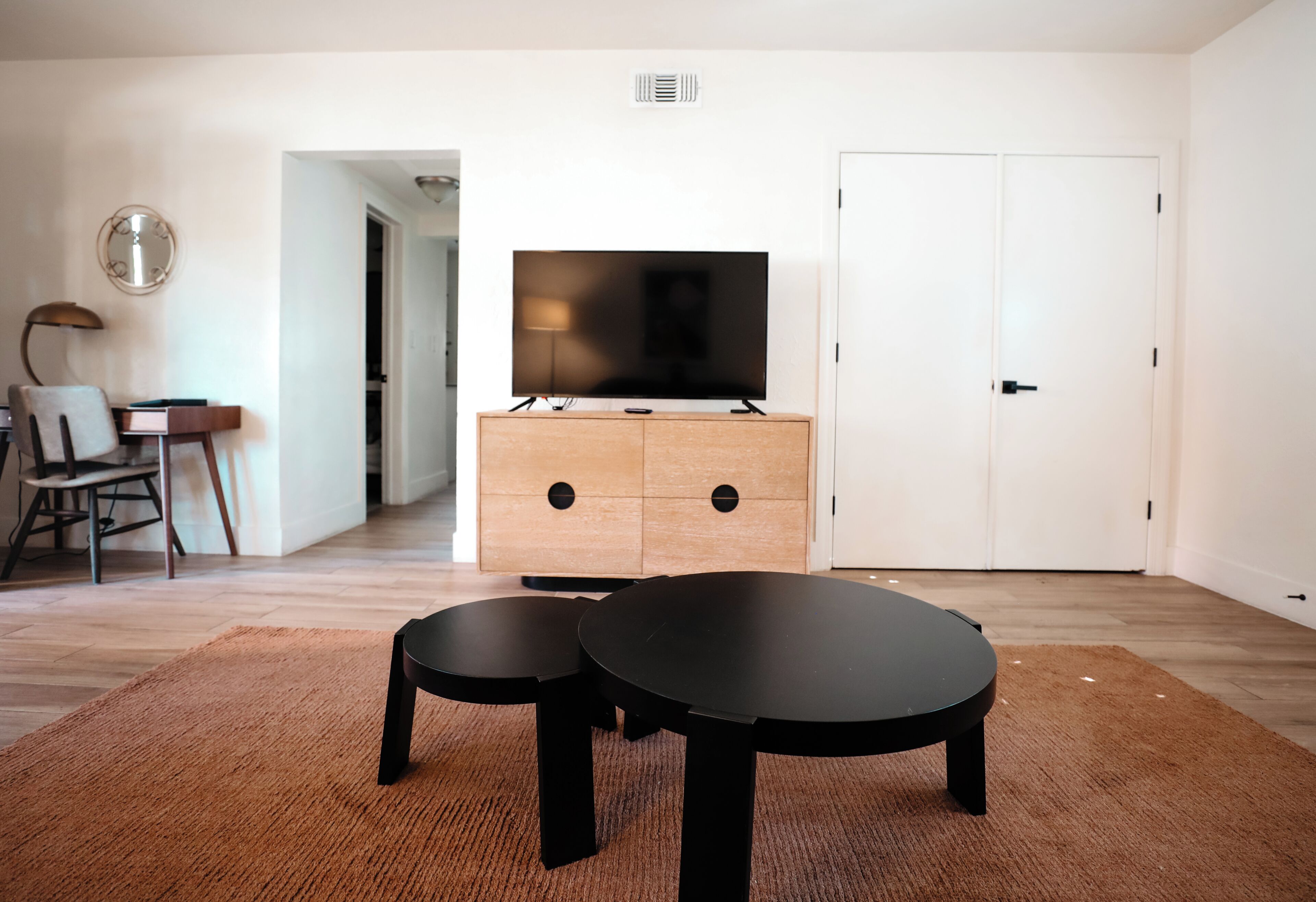A modern living room with a flat-screen TV on a wooden cabinet, two black round tables, a brown rug, a chair, and a lamp.