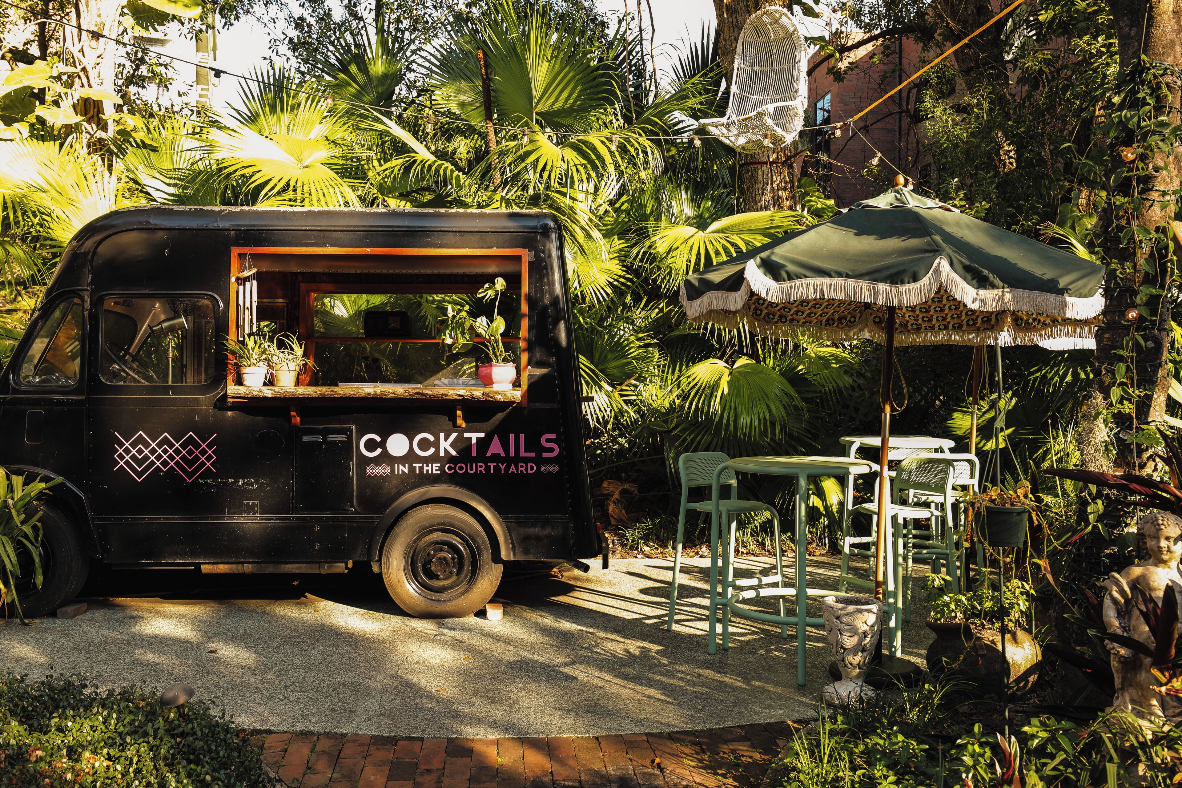 A black food truck with 'COCKTAILS IN THE COURTYARD' written on it, parked in a garden with a table and chairs under a canopy.