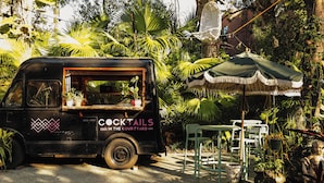 Uma food truck preta com a inscrição 'COCKTAILS IN THE COURTYARD', estacionada em um jardim com uma mesa e cadeiras sob um toldo.
