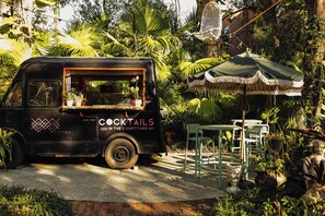 A black food truck with 'COCKTAILS IN THE COURTYARD' written on it, parked in a garden with a table and chairs under a canopy.