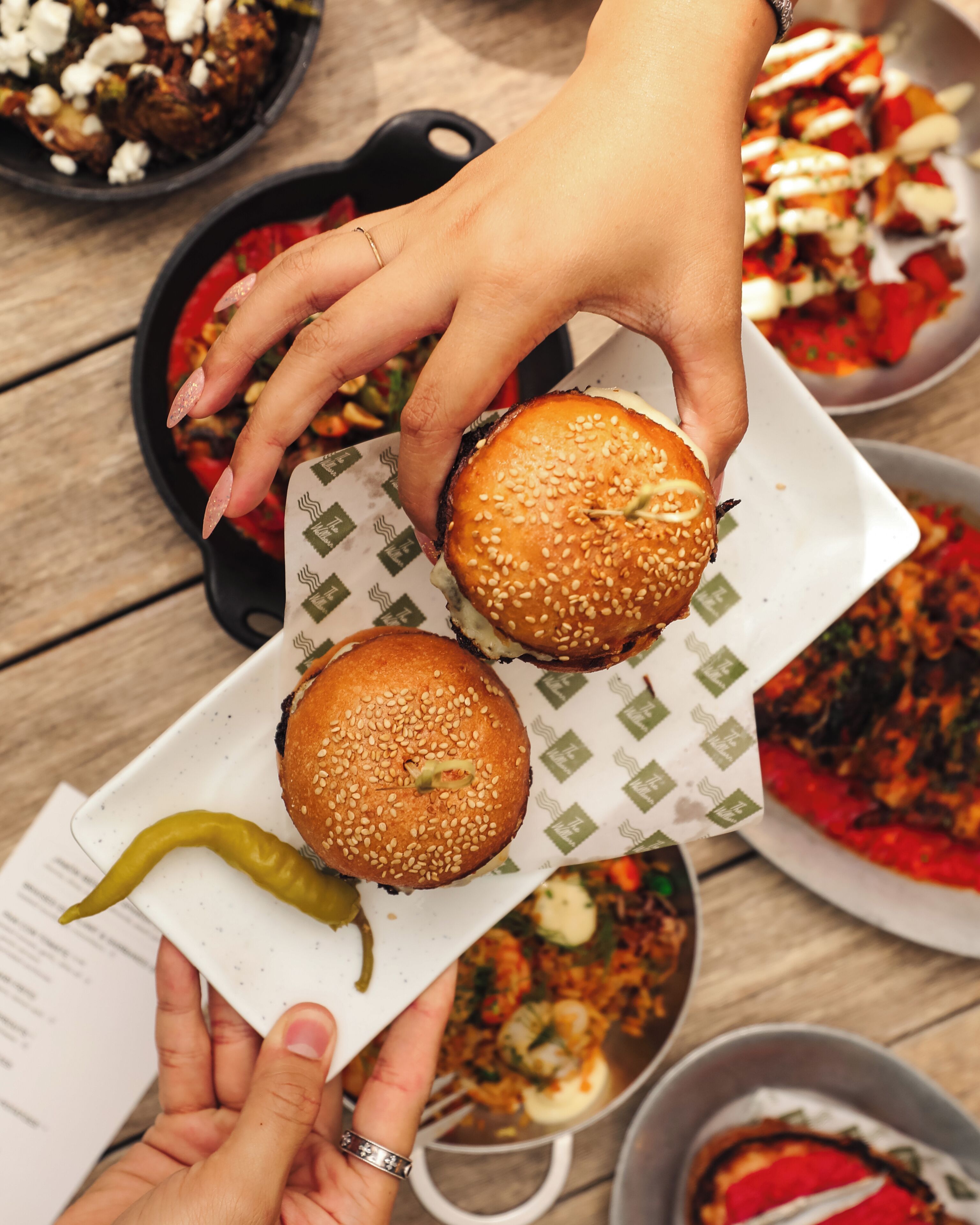 Une personne tenant deux hamburgers garnis de graines de sésame sur une assiette blanche à motifs verts, avec un piment vert et un menu à proximité.