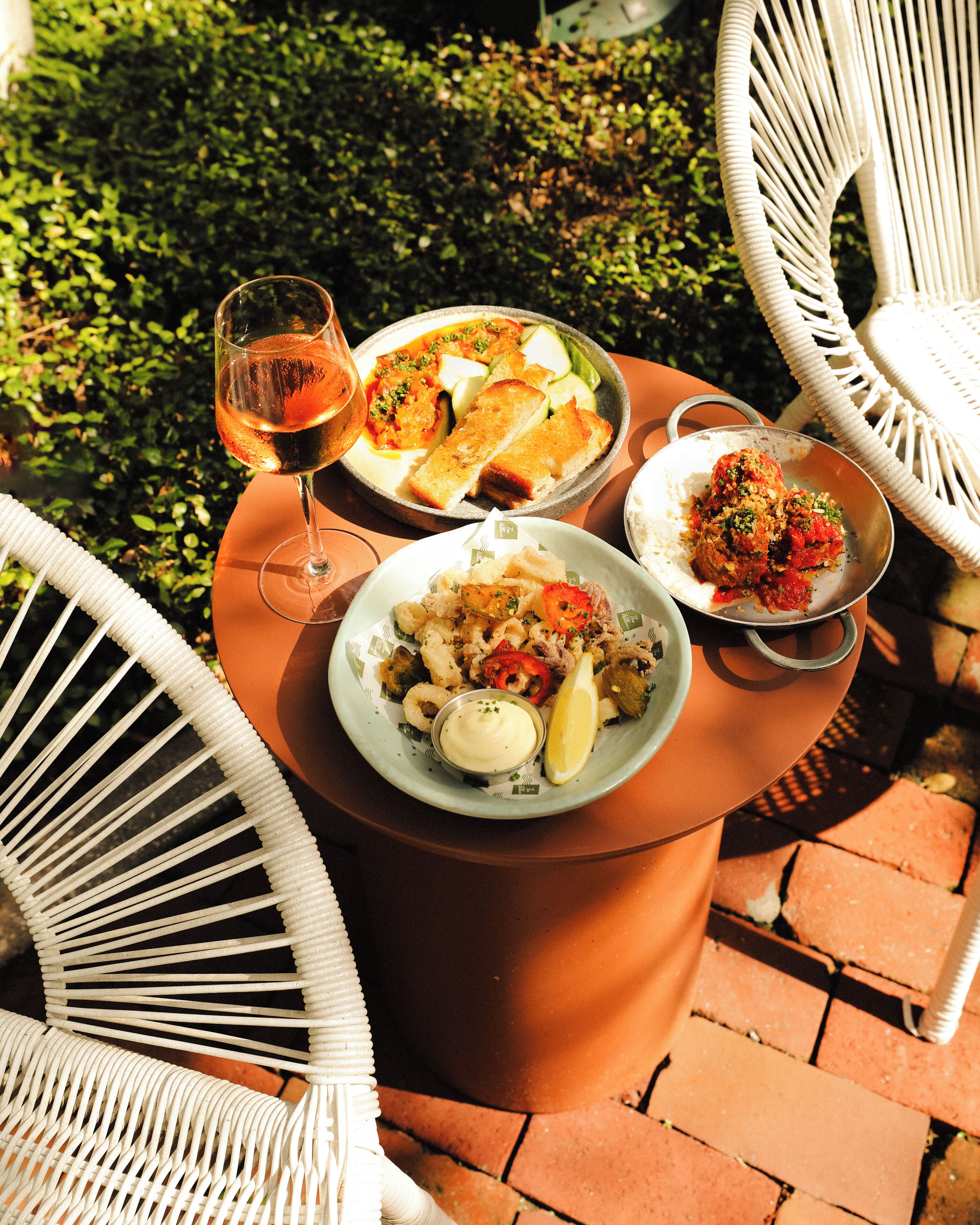 A table with plates of food and a glass of wine, surrounded by white chairs.
