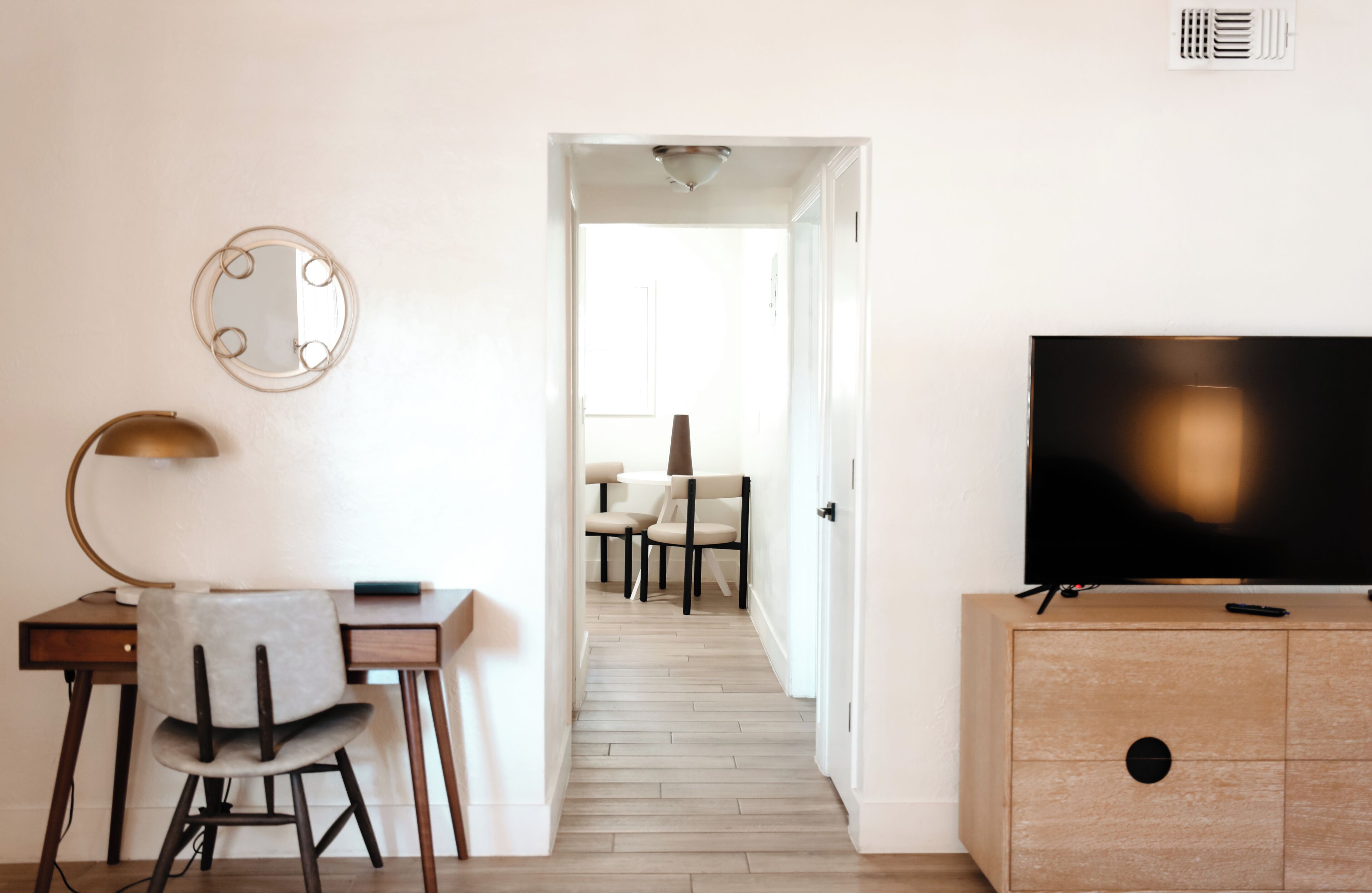 A modern hotel room with a desk, chair, television, and a hallway leading to a dining area.