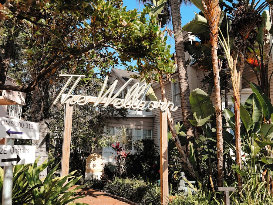 A sign with 'The Wellborn' written on it, surrounded by lush greenery and a building in the background.