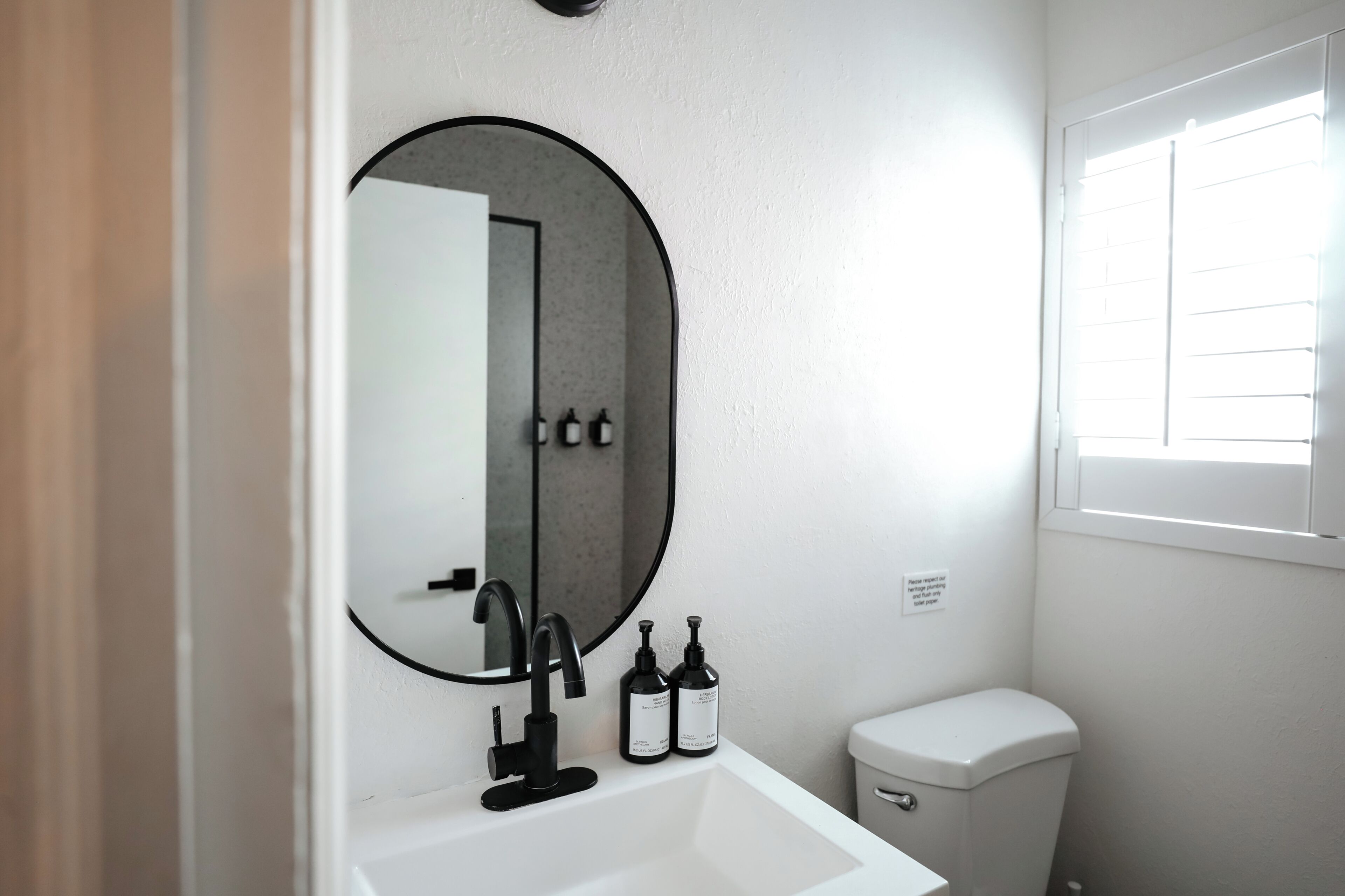 A bathroom with a white sink, black faucet, and a round mirror. There is a toilet to the right and a window above it.