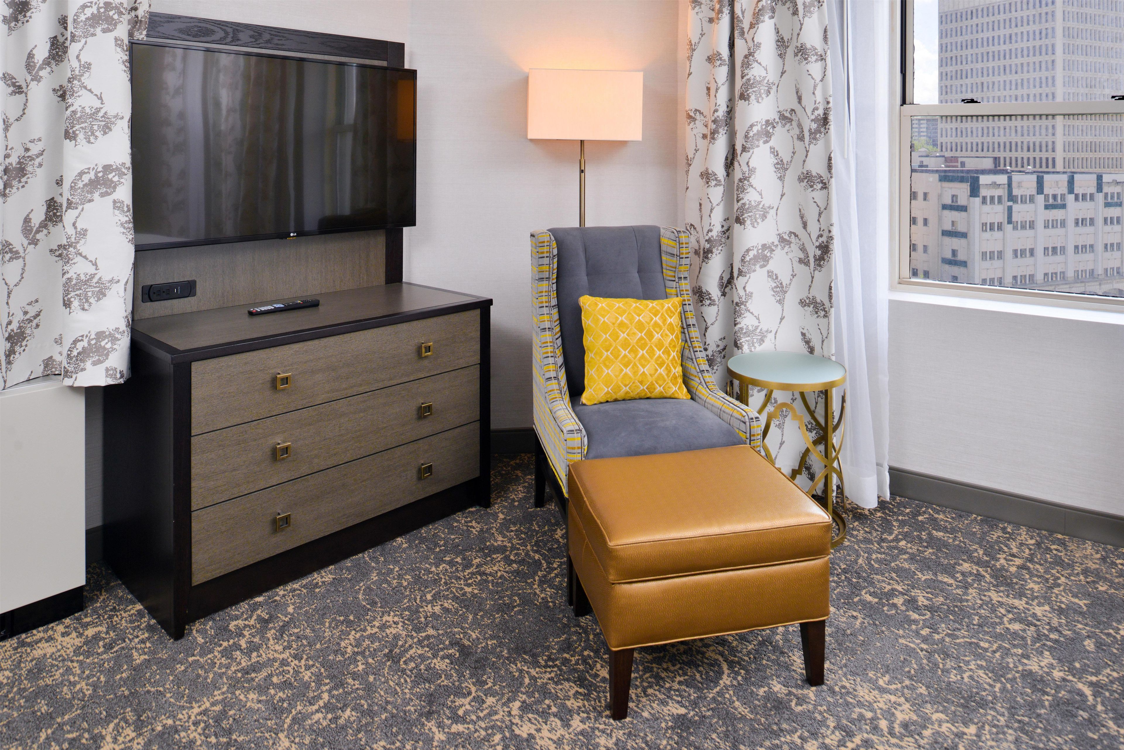 A hotel room with a flat-screen TV, a desk with drawers, a chair with a cushion, a small round table, and a window with a view of buildings.