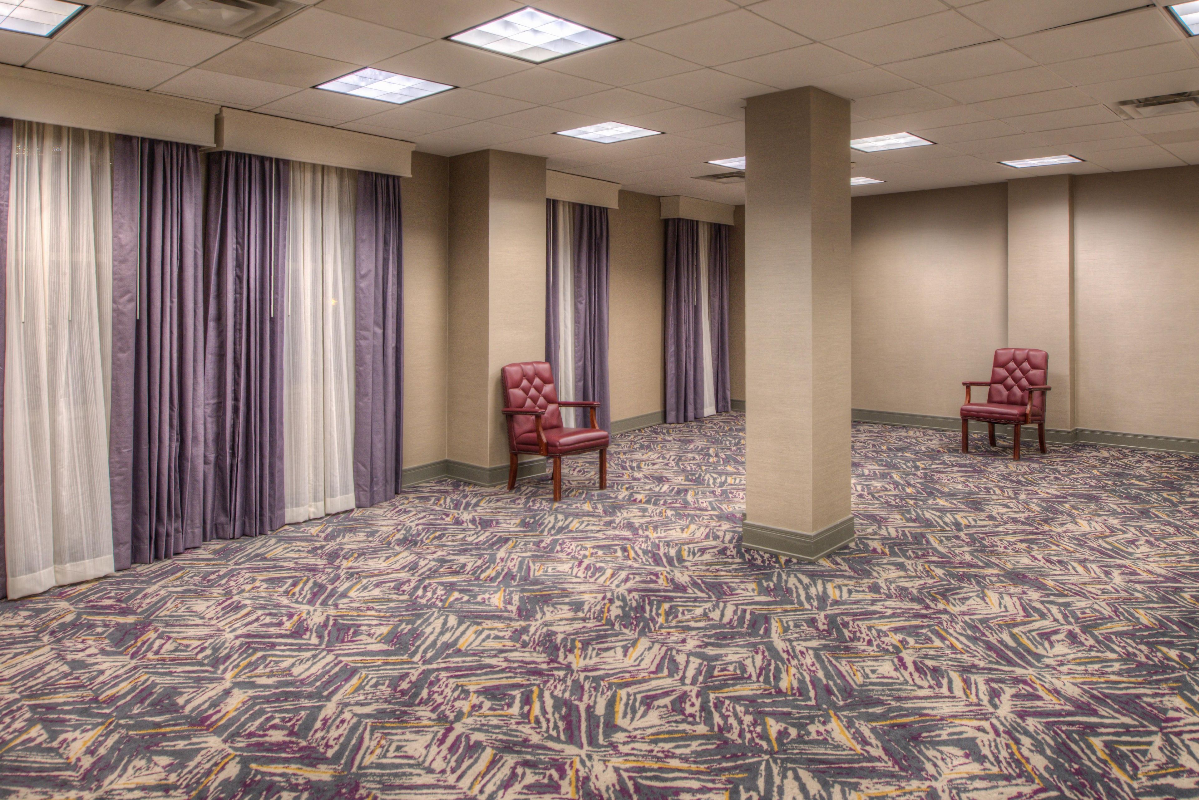 A hotel room with a patterned carpet, a large window with curtains, and two red upholstered chairs.