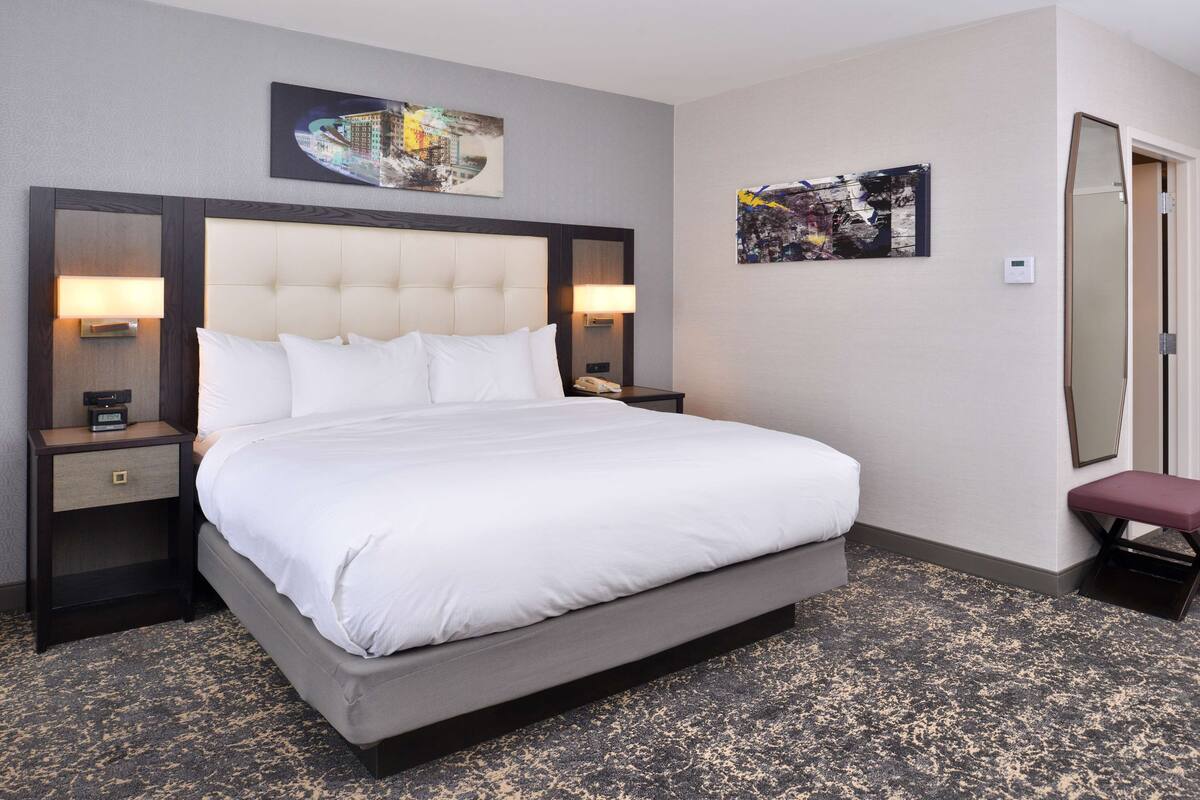 A hotel room with a large bed, two bedside tables with lamps, a wall-mounted artwork, and a bench by the door.