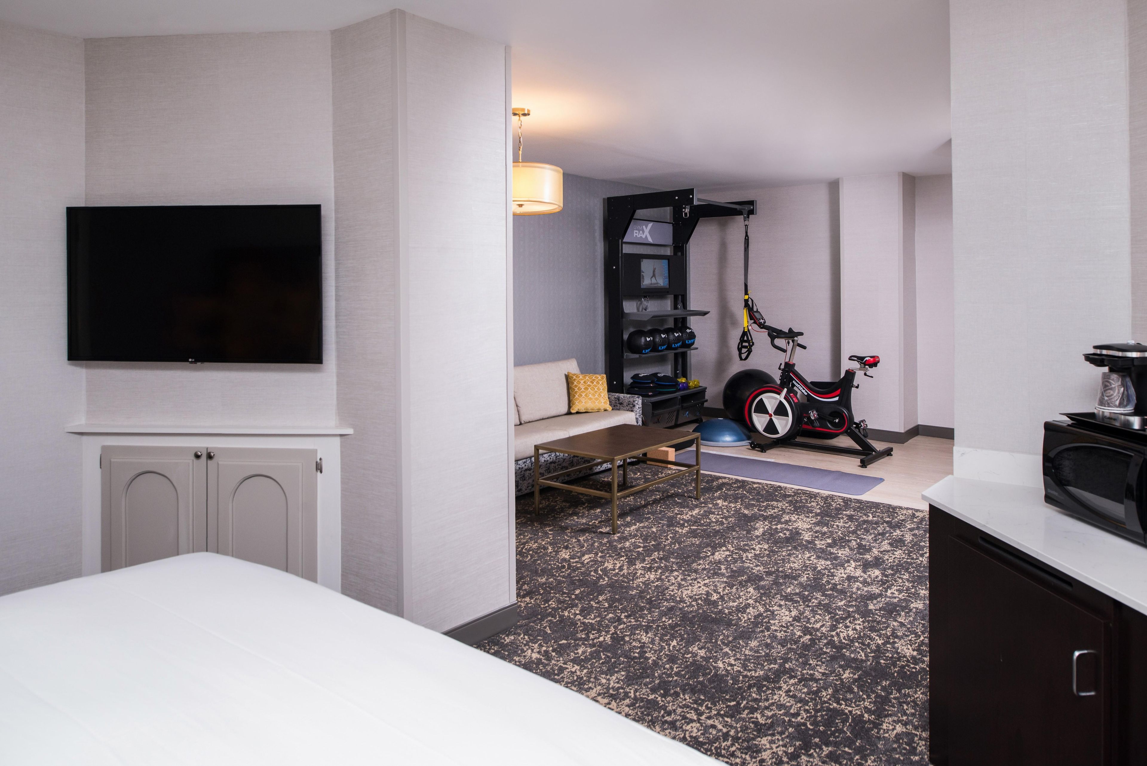 Ein Hotelzimmer mit einem an der Wand befestigten Flachbildfernseher, einem Sofa, einem Couchtisch, einem Laufband und einem kleinen Gymnastikball.