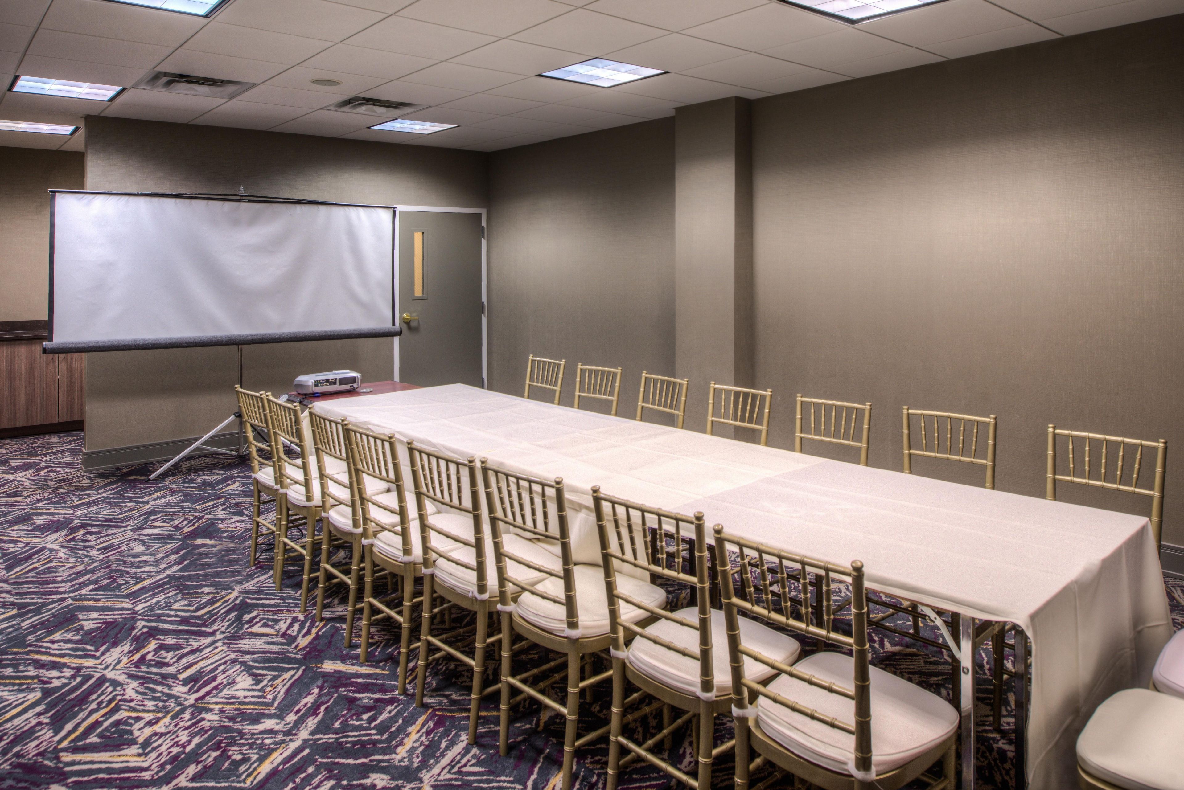 A conference room with a long table, chairs, a projector screen, and a carpeted floor.