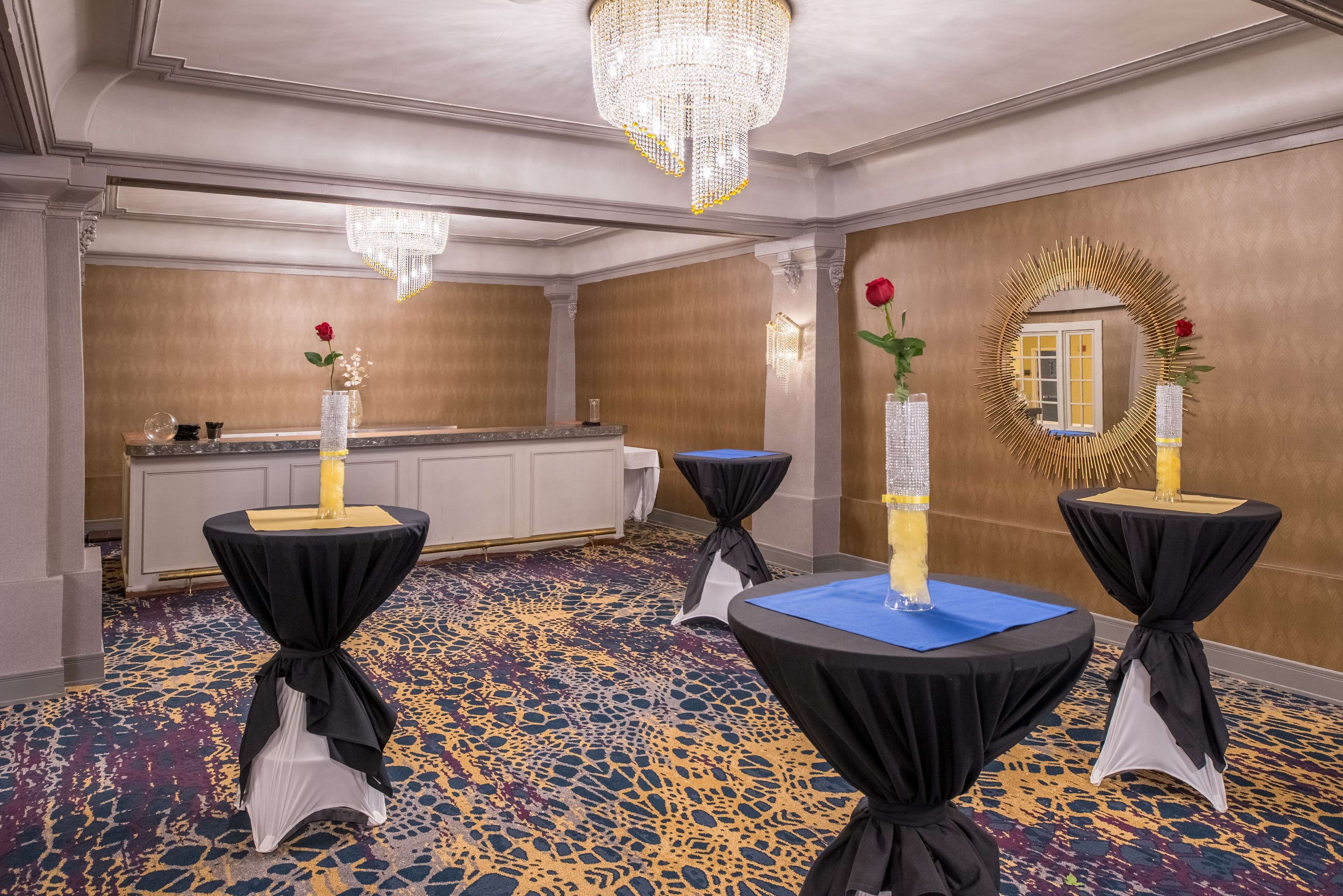 A room with a chandelier, round tables with black and white tablecloths, a mirror with a gold frame, and a patterned carpet.