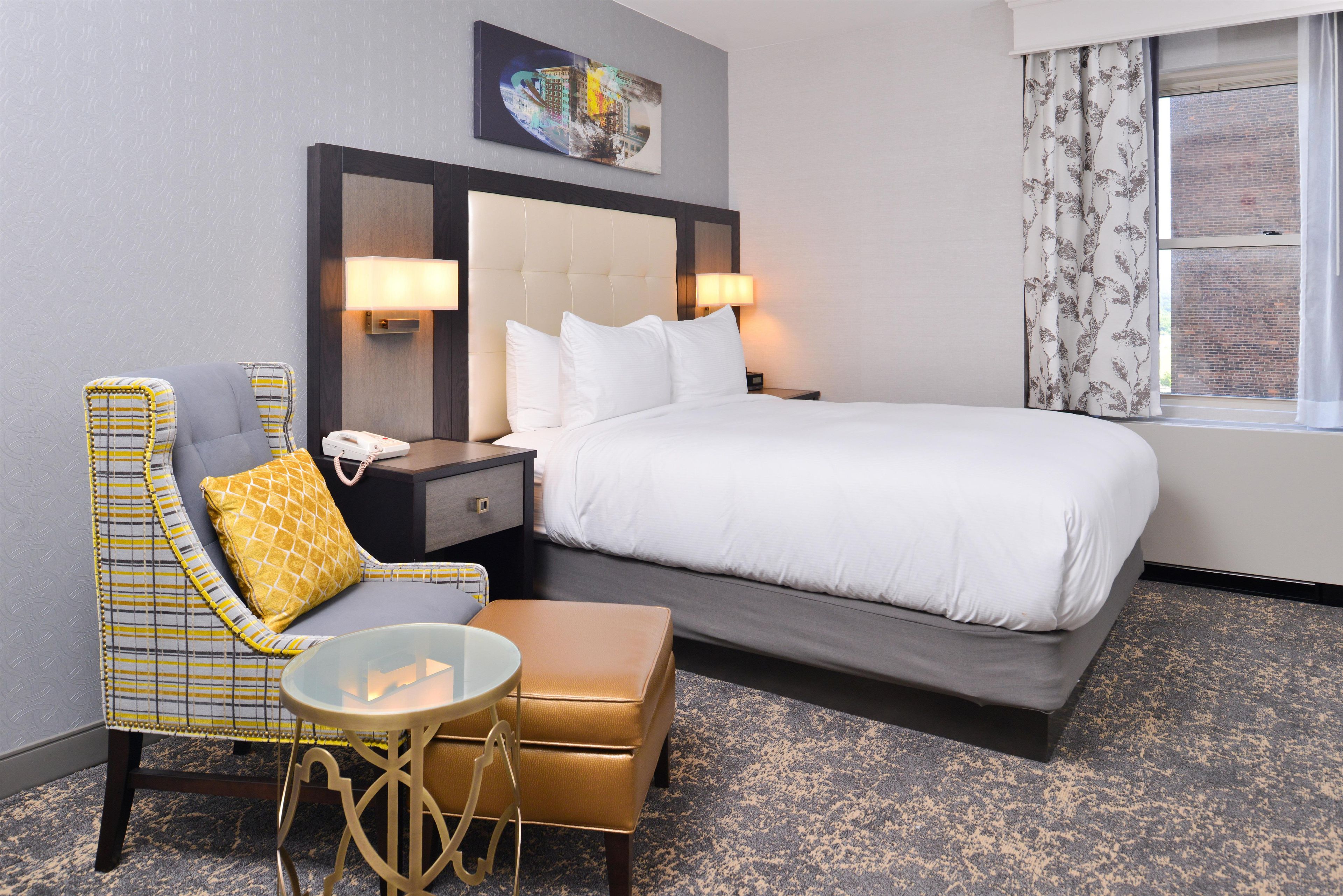 A hotel room with a bed, a chair, a small table, a nightstand, and a window with curtains.