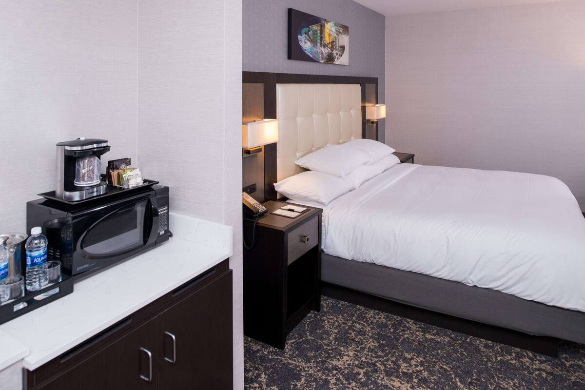 A hotel room with a bed, a nightstand with a coffee maker, a microwave, and a small table with water bottles.