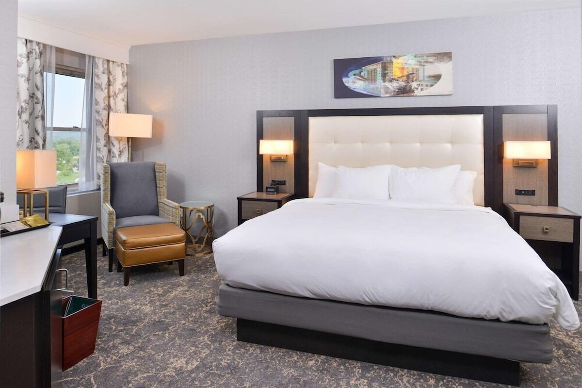 Ein Hotelzimmer mit einem großen Bett, zwei Nachttischlampen, einem Schreibtisch mit Stuhl, einem kleinen Hocker und einem Fenster mit Vorhängen.