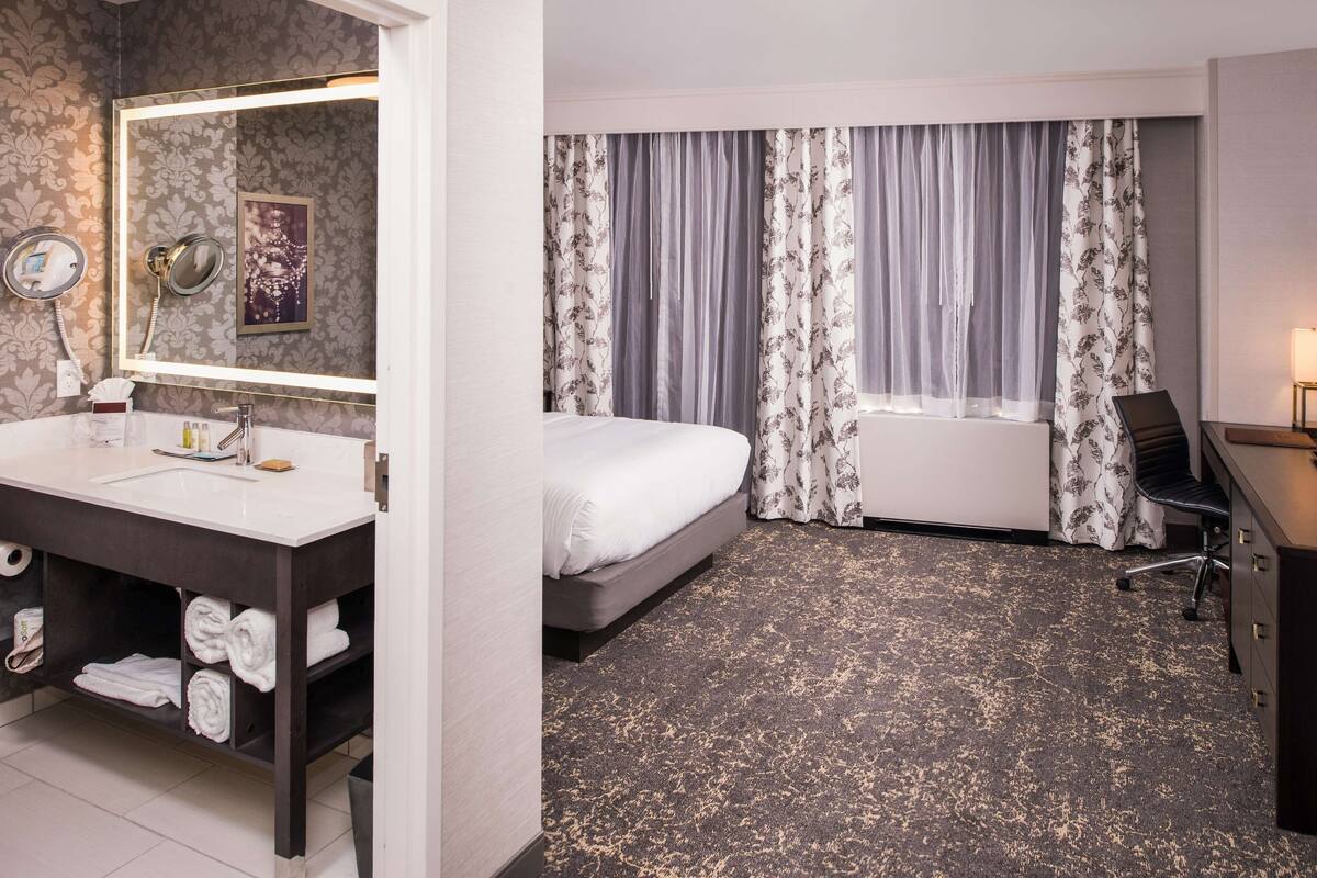 A hotel room with a large bed, a bathroom with a bathtub, a vanity with a mirror, and a desk with a chair.