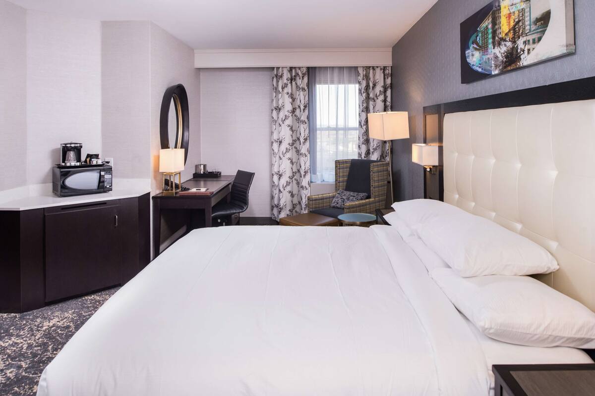 A hotel room with a large bed, a desk with a computer, a chair, a small table, and a lamp.