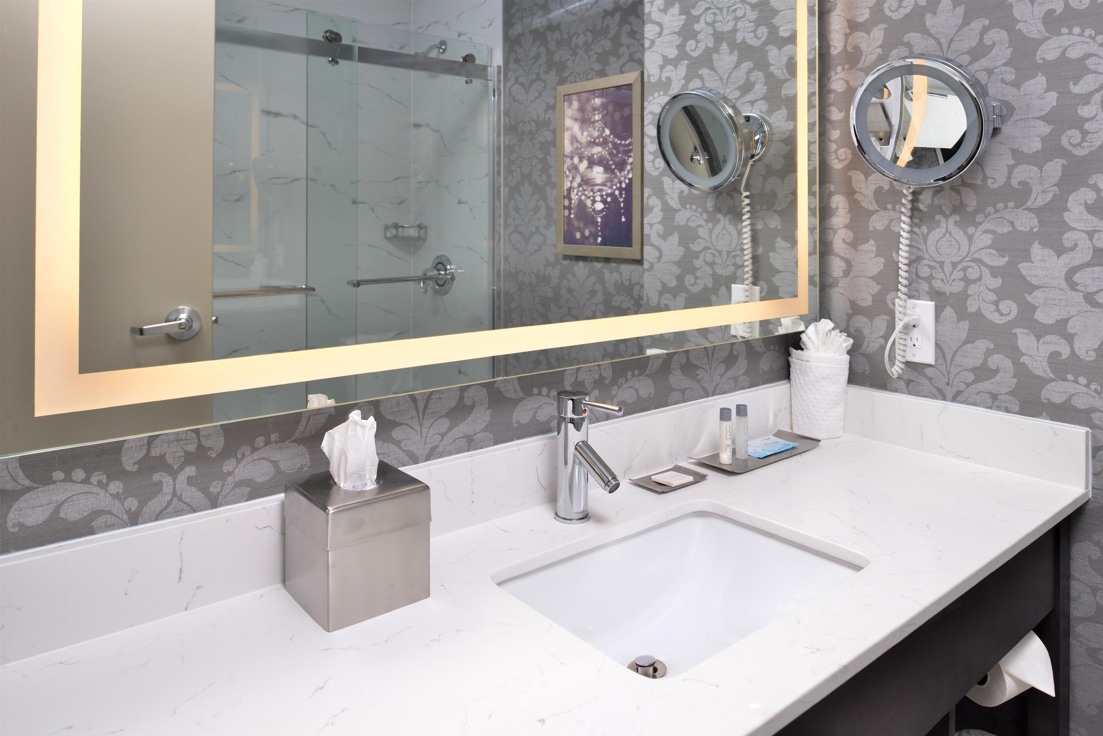 Une salle de bain dotée d’un grand miroir, d’un lavabo, d’un distributeur de serviettes et d’une cabine de douche en verre.