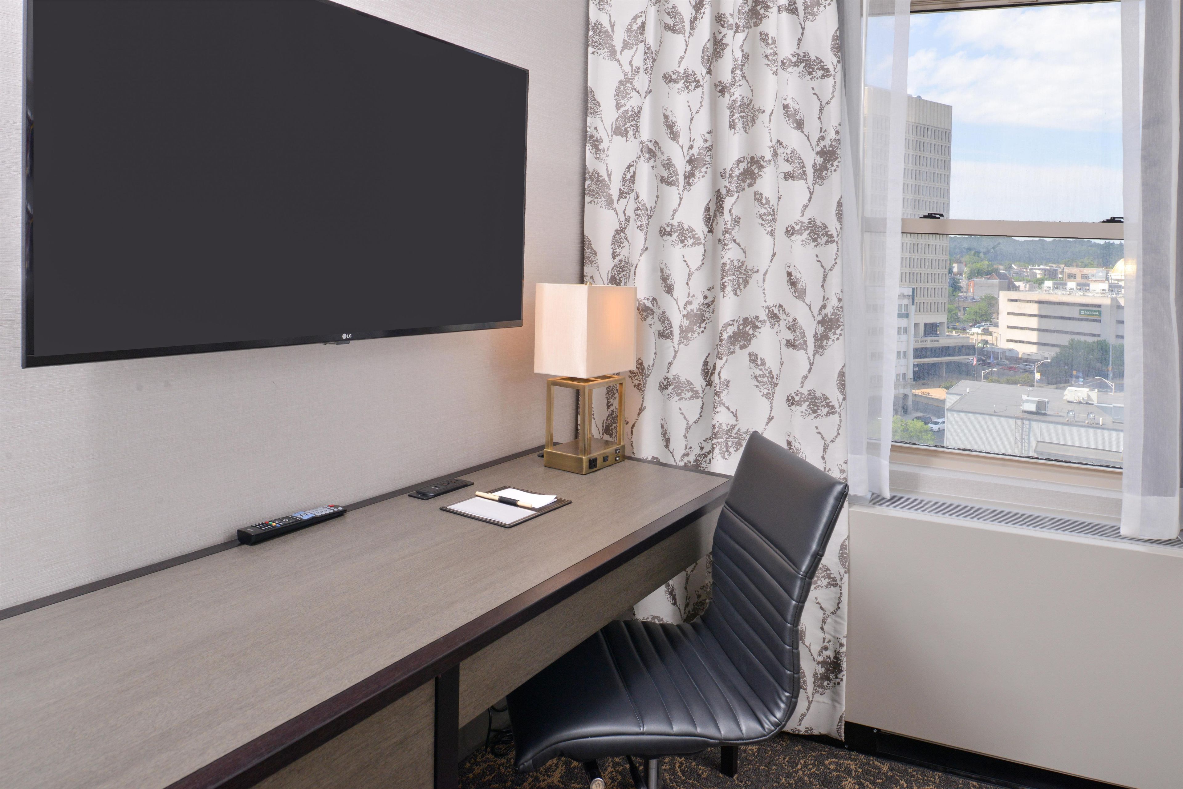 Habitación de hotel con una televisión de pantalla plana montada en la pared, un escritorio con lámpara, una silla y una ventana con vistas a la ciudad.