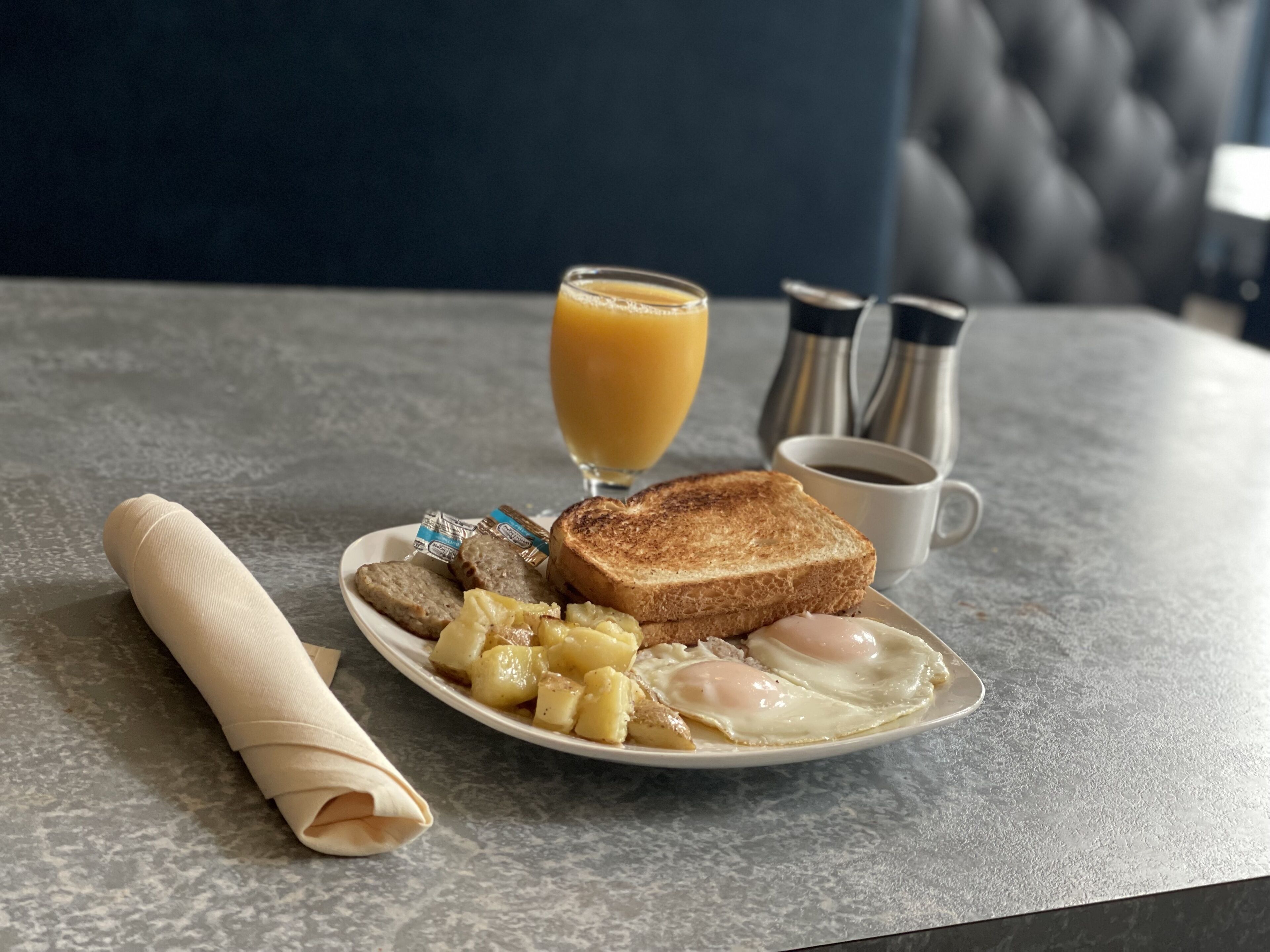 Un plato de desayuno con tostadas, huevos, salchichas y papas, un vaso de jugo de naranja, una taza de café y una servilleta doblada sobre una mesa.