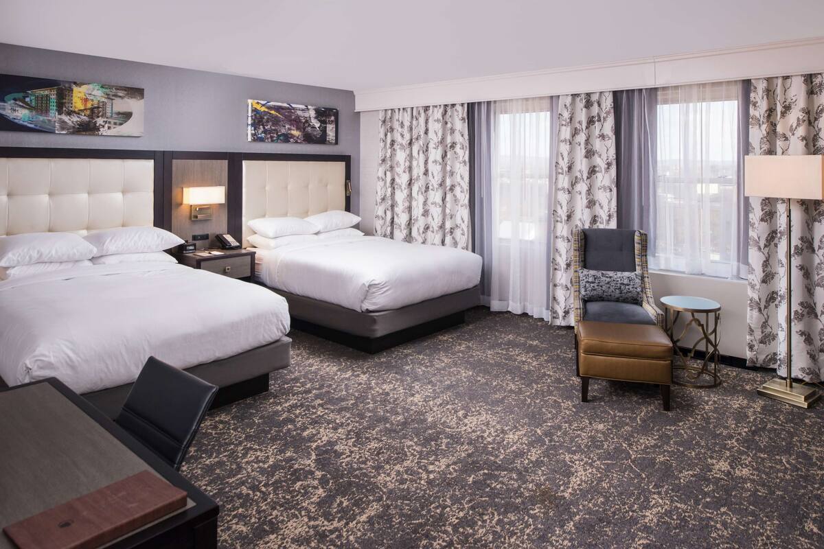 A hotel room with two beds, a desk, a chair, a small table, and a lamp.