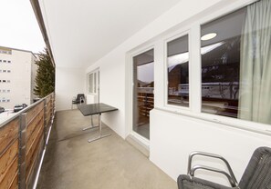 Un balcón con mesa y sillas, vista a un edificio y una ventana con vistas a un paisaje nevado.