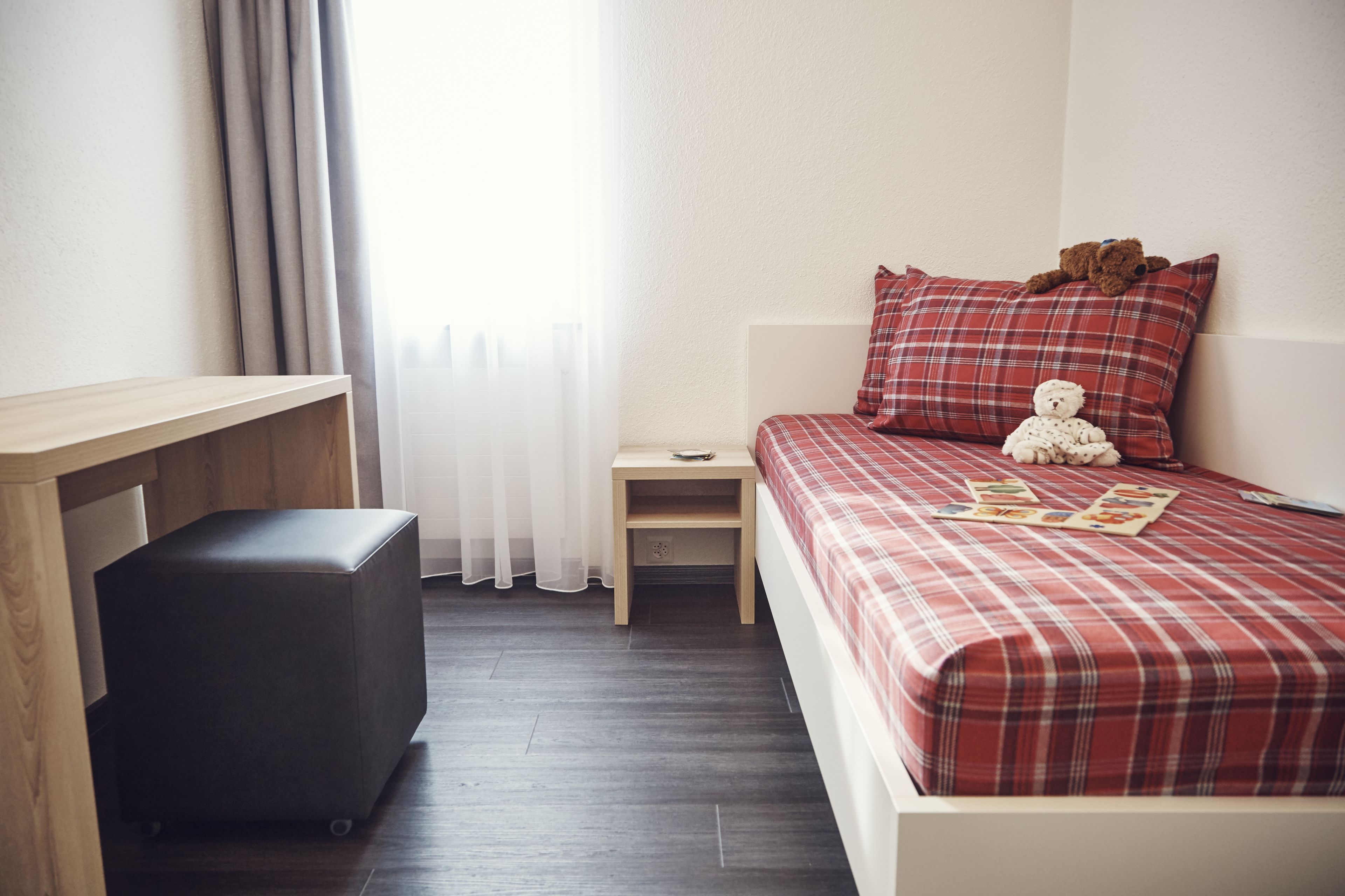 Um quarto com cama, mesa de cabeceira, escrivaninha e um pufe para guardar objetos.