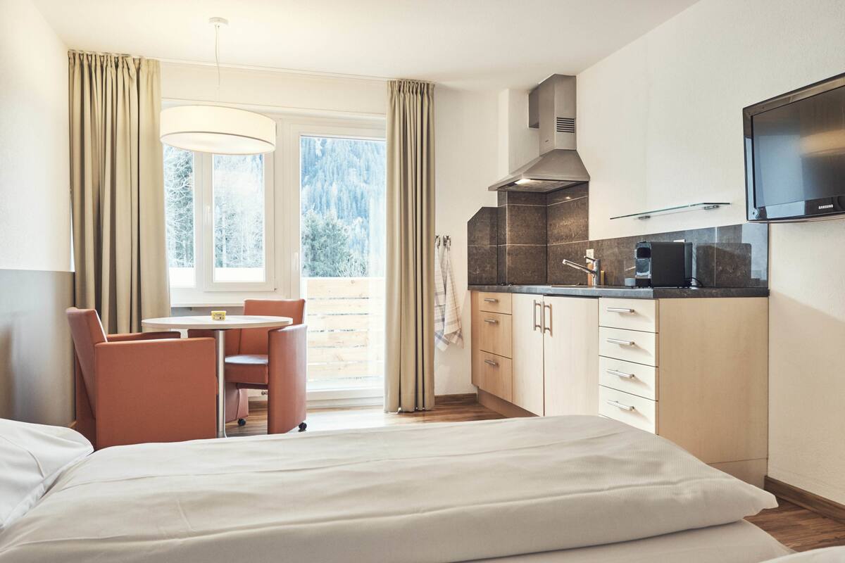 Habitación de hotel con cama, una mesa de comedor con sillas, kitchenette y un ventanal con cortinas.