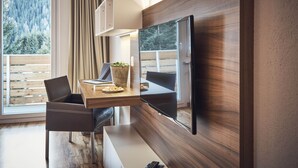 Una habitación moderna con televisor de pantalla plana en la pared, una mesa de comedor de madera con sillas, y vistas a una terraza de madera y árboles a través de un ventanal grande.