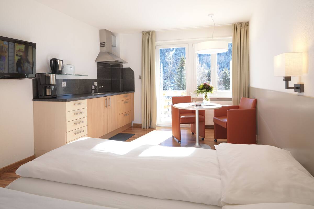 Habitación de hotel con cama, una pequeña mesa de comedor con sillas, una zona de cocina con cafetera y ventana con cortinas.
