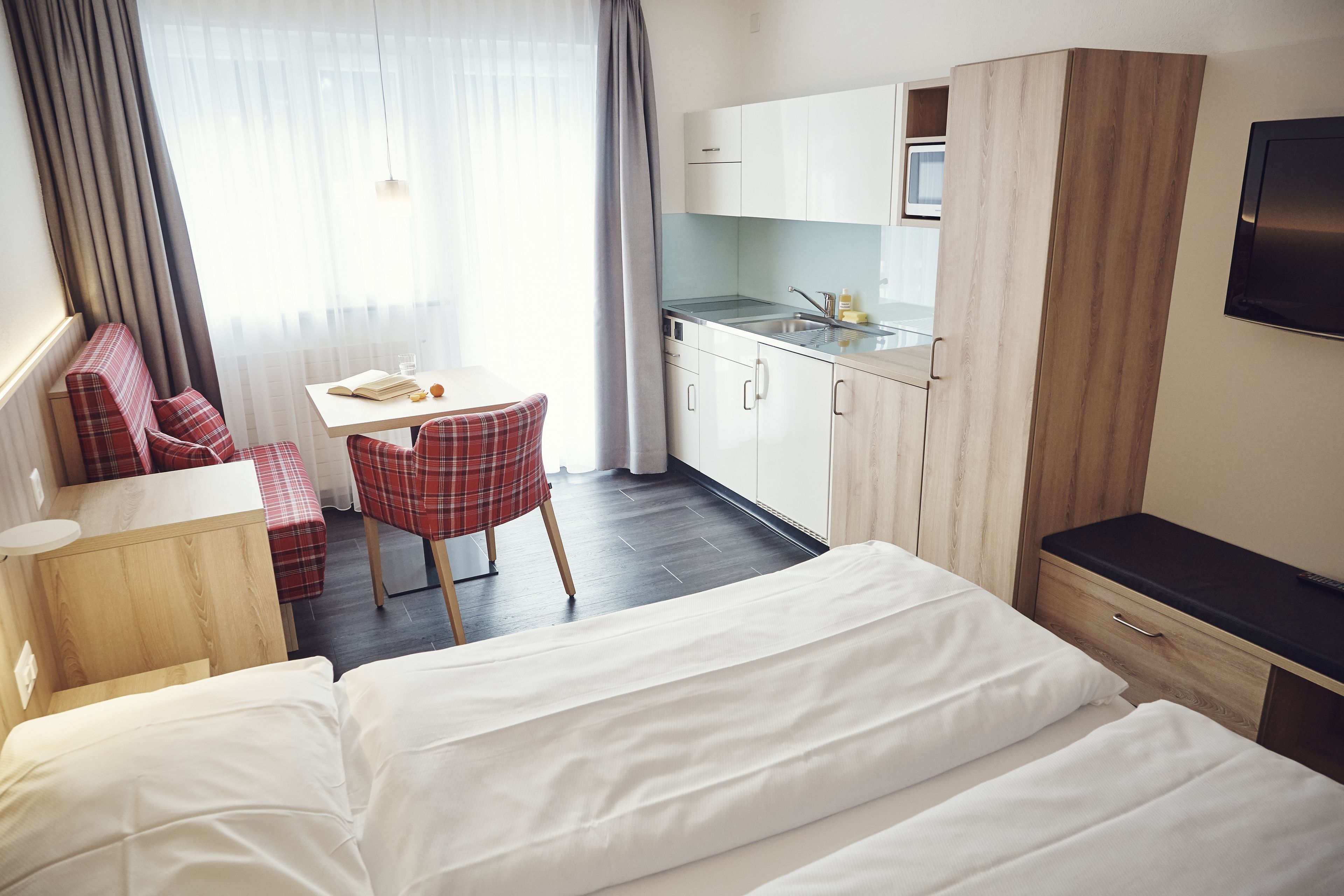 A hotel room with a bed, a desk, a chair, a small table, a kitchenette, and a TV.