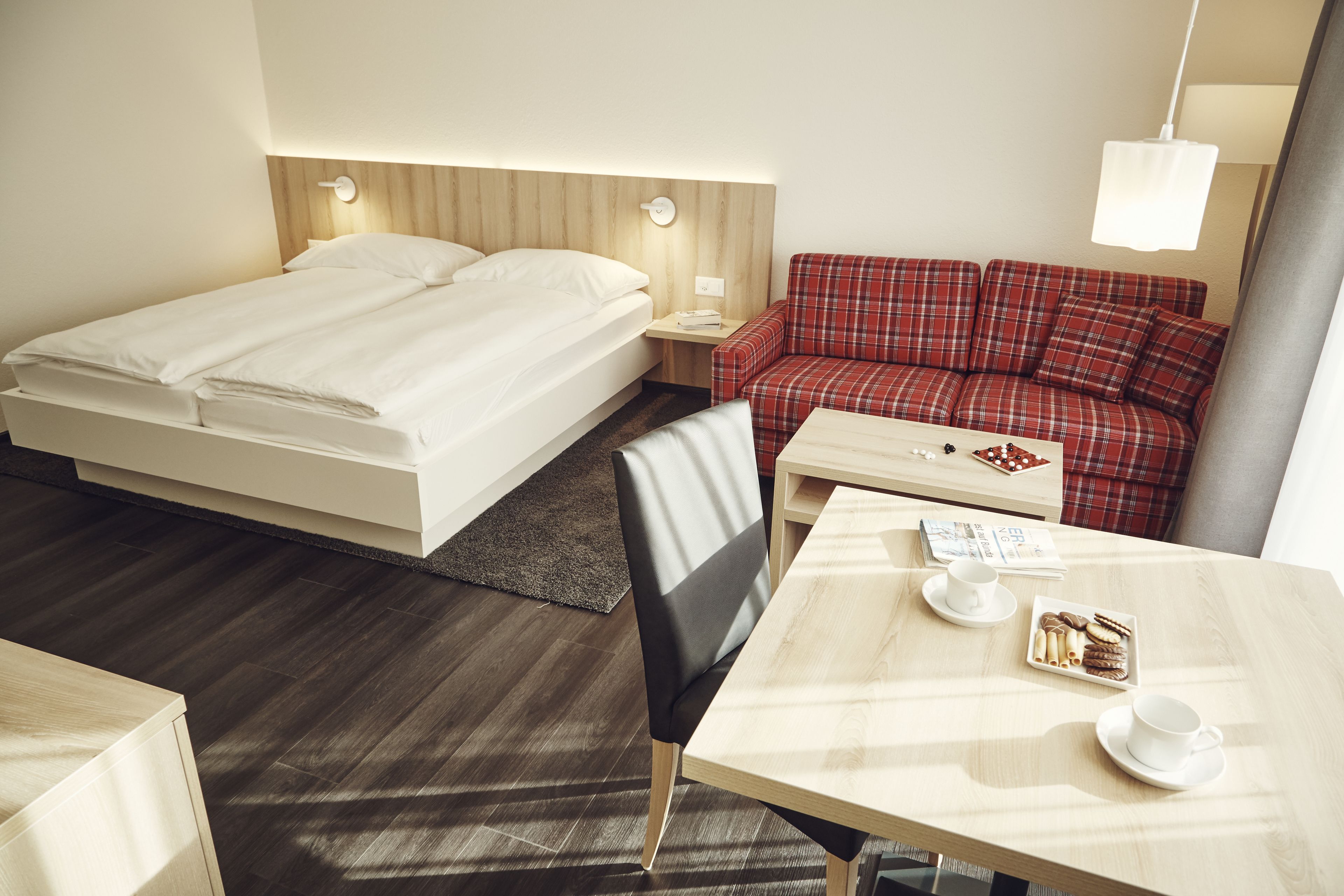A hotel room with a bed, a red plaid sofa, a wooden table with a tray of cookies and a cup, and a nightstand with a lamp.