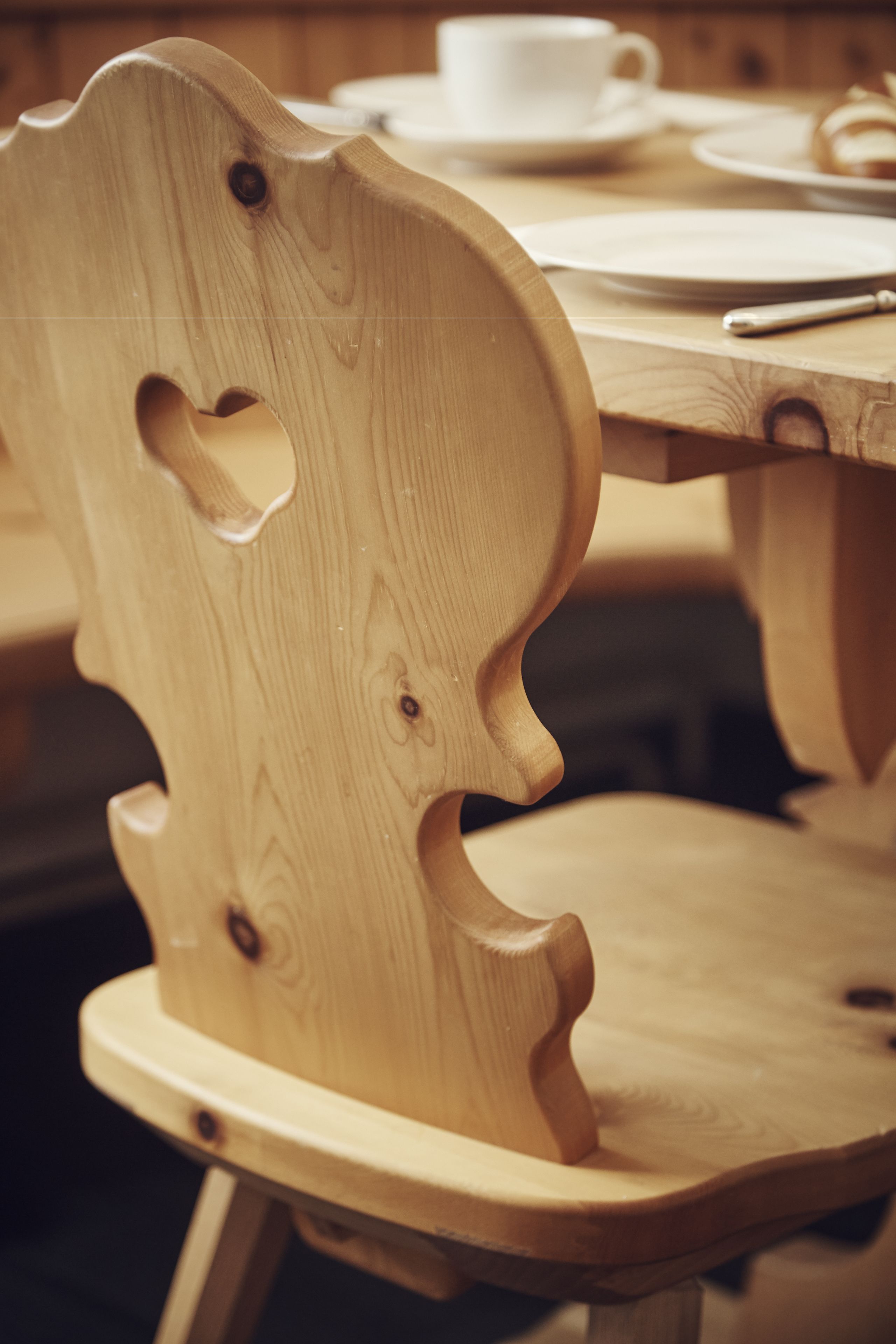 A wooden chair with a unique cutout design, a white cup, and a plate on a wooden table.
