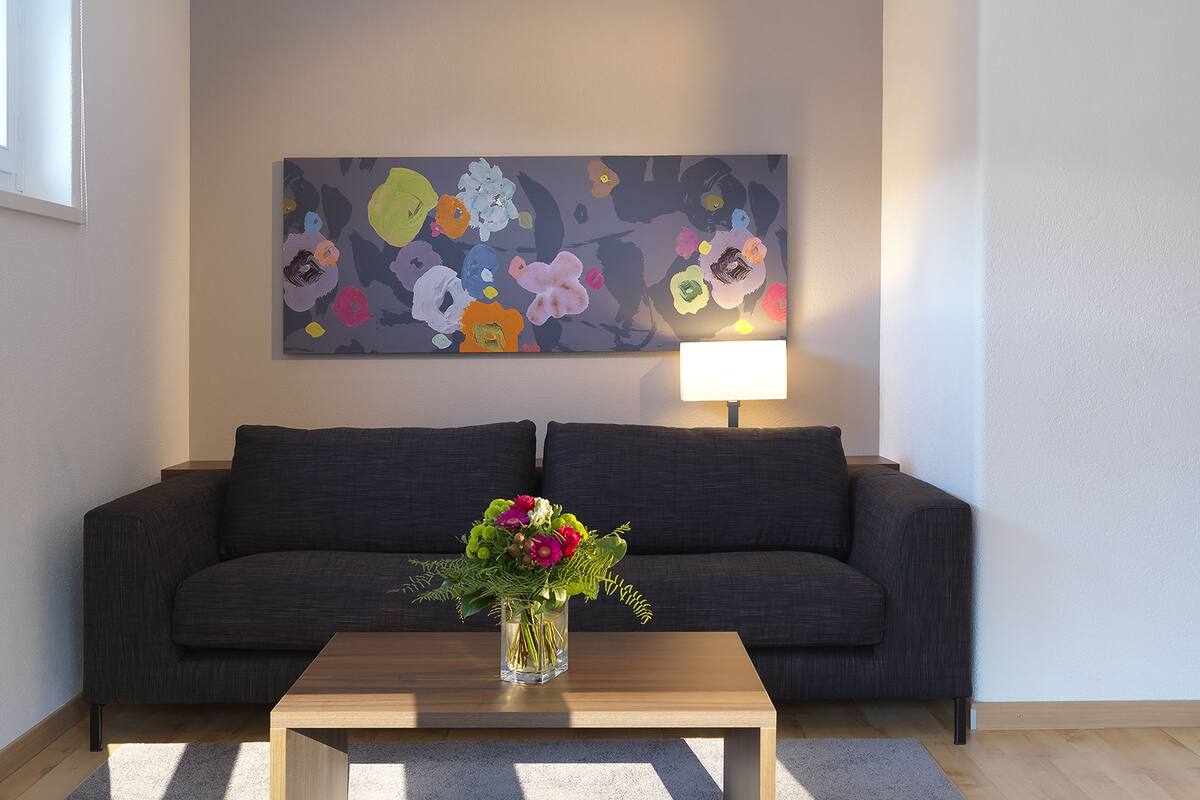 Una sala de estar con un sofá de color oscuro, una mesa de centro de madera con un jarrón de flores y un cuadro abstracto y colorido en la pared.