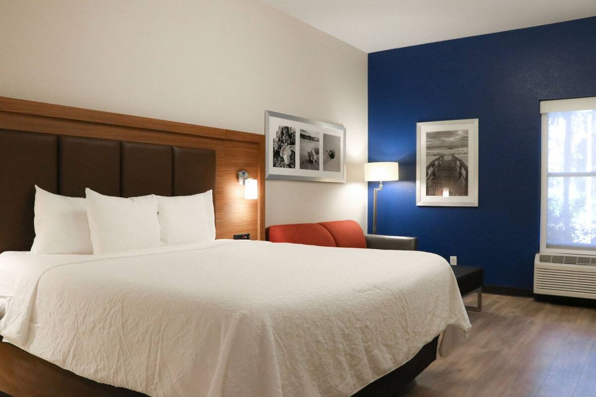 Une chambre d’hôtel avec un grand lit, une tête de lit en bois, une chaise rouge, une table de chevet, une lampe et deux tableaux encadrés au mur.