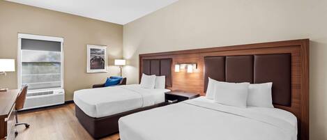 A hotel room with two beds, a desk, a chair, a window with blinds, and a wall-mounted lamp.