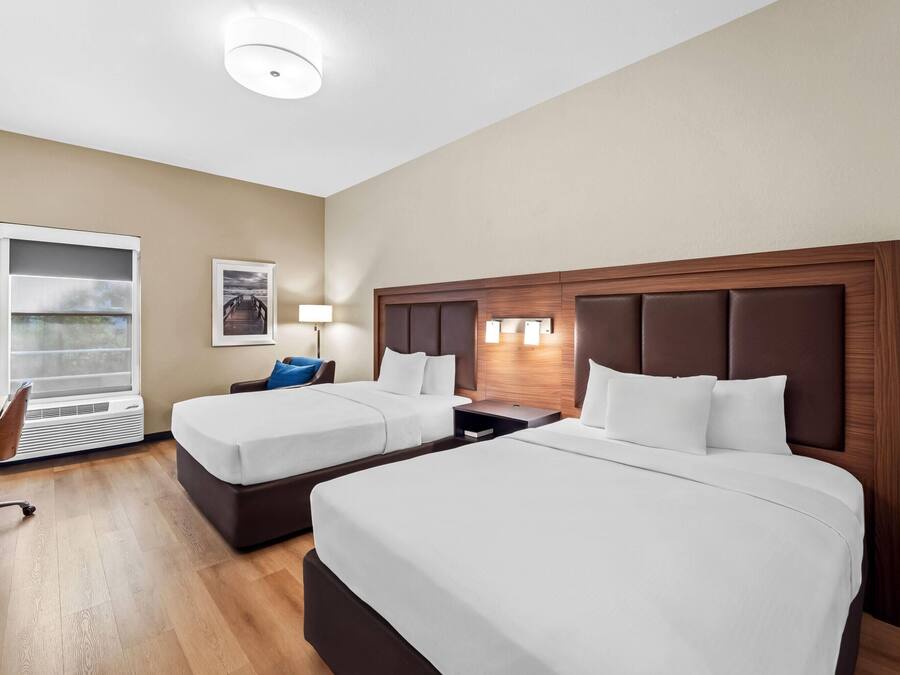 A hotel room with two beds, a desk, a chair, a window with blinds, and a wall-mounted lamp.