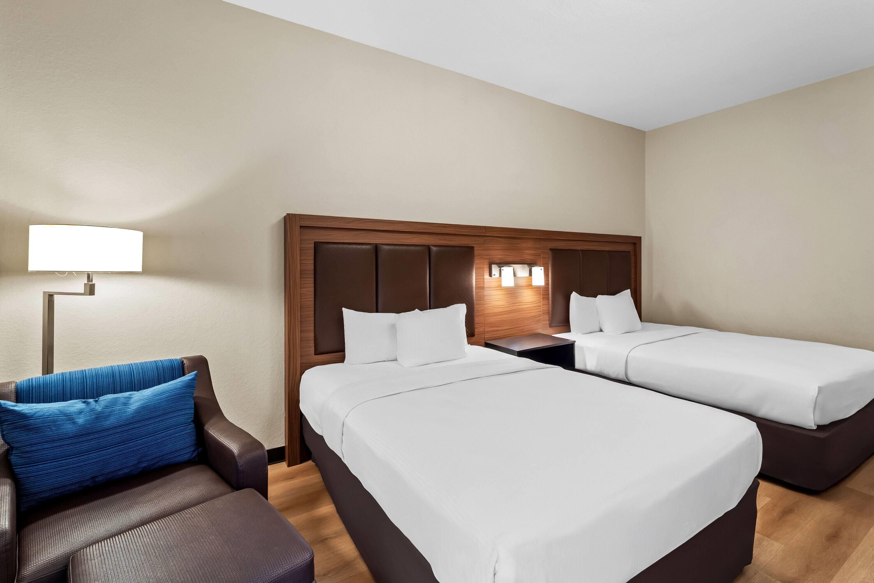 Habitación de hotel con dos camas individuales, un sofá, una mesita de noche y una lámpara.