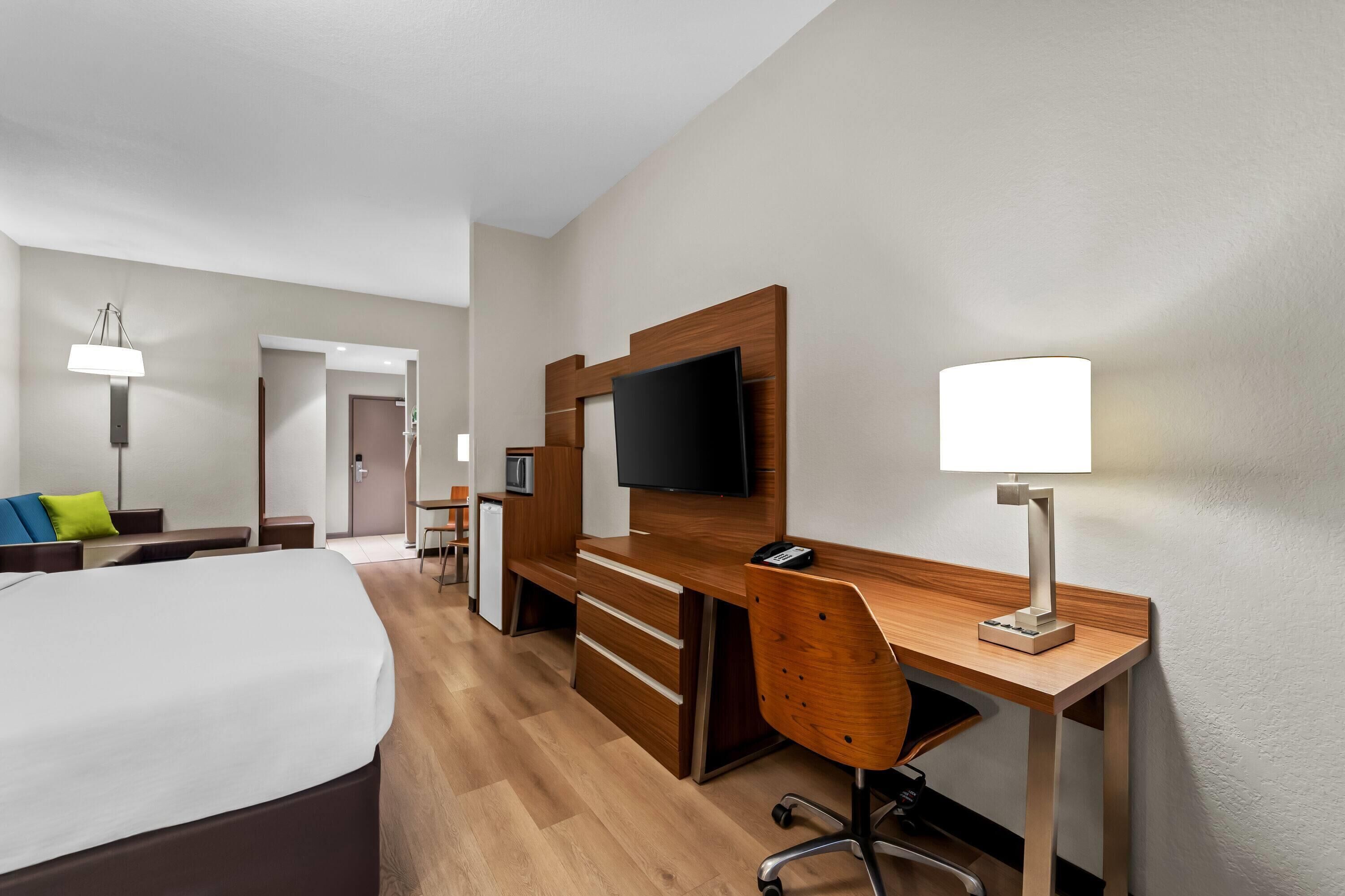 A hotel room with a large bed, a wooden desk with a TV, a chair, and a desk lamp.