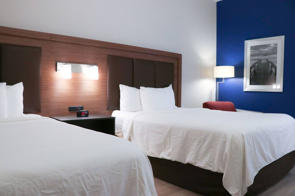 Une chambre d’hôtel avec deux lits, une table de chevet avec une horloge, une chaise rouge et un tableau encadré au mur.